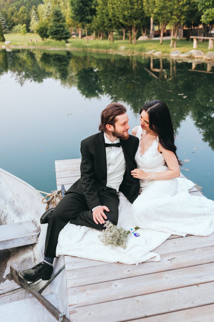 A romantic wedding couple sits on a wooden dock by a serene lake surrounded by trees.