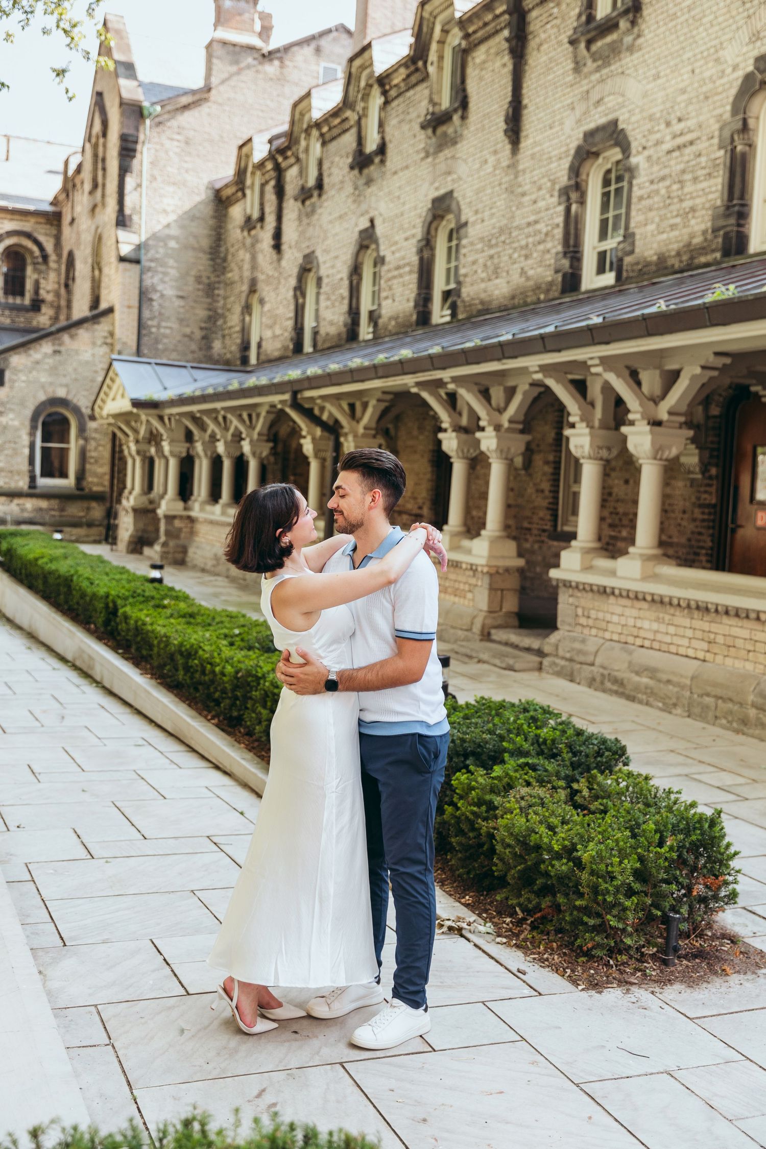 A couple embraces in front of a historic stone building with Gothic architectural features and manicured gardens.