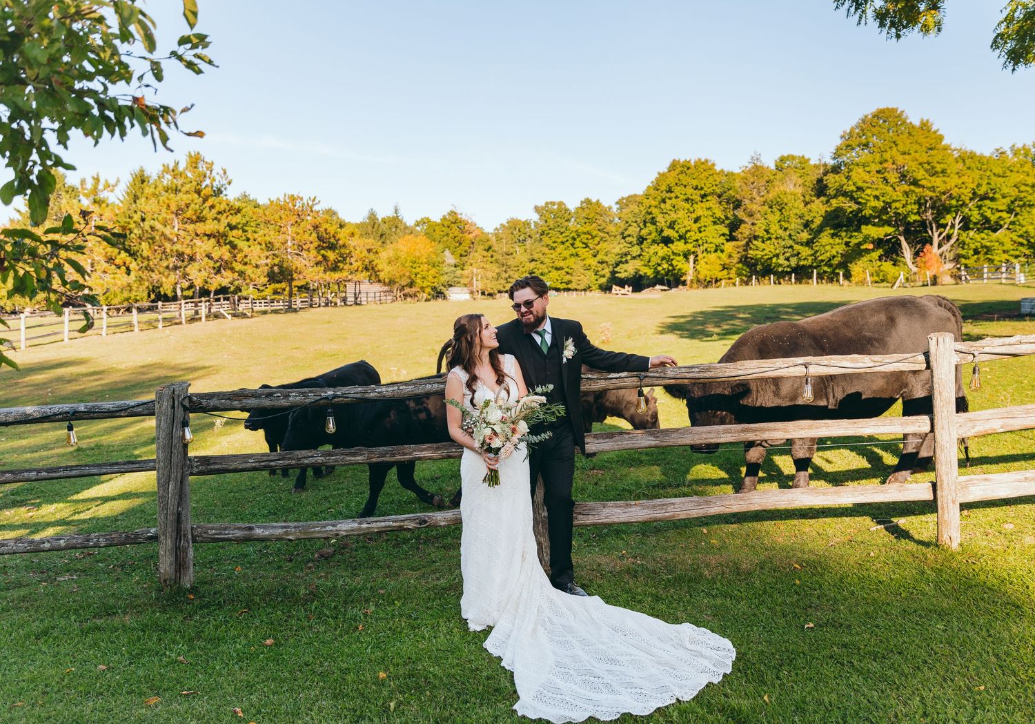 A couple embraces in front of a rustic wooden fence during sunset in a pastoral setting.