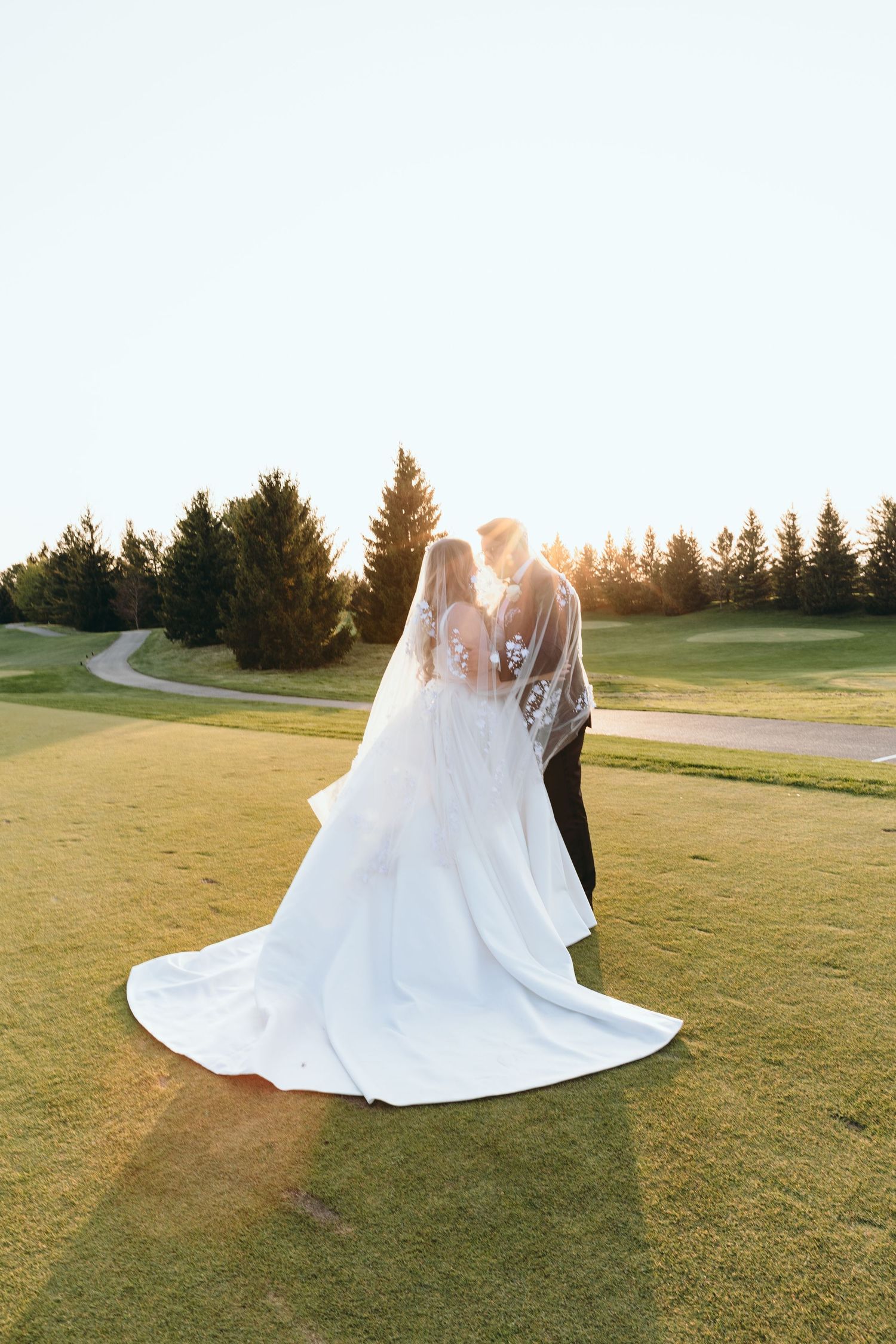 A couple in wedding attire share an intimate moment on a golf course at sunset.
