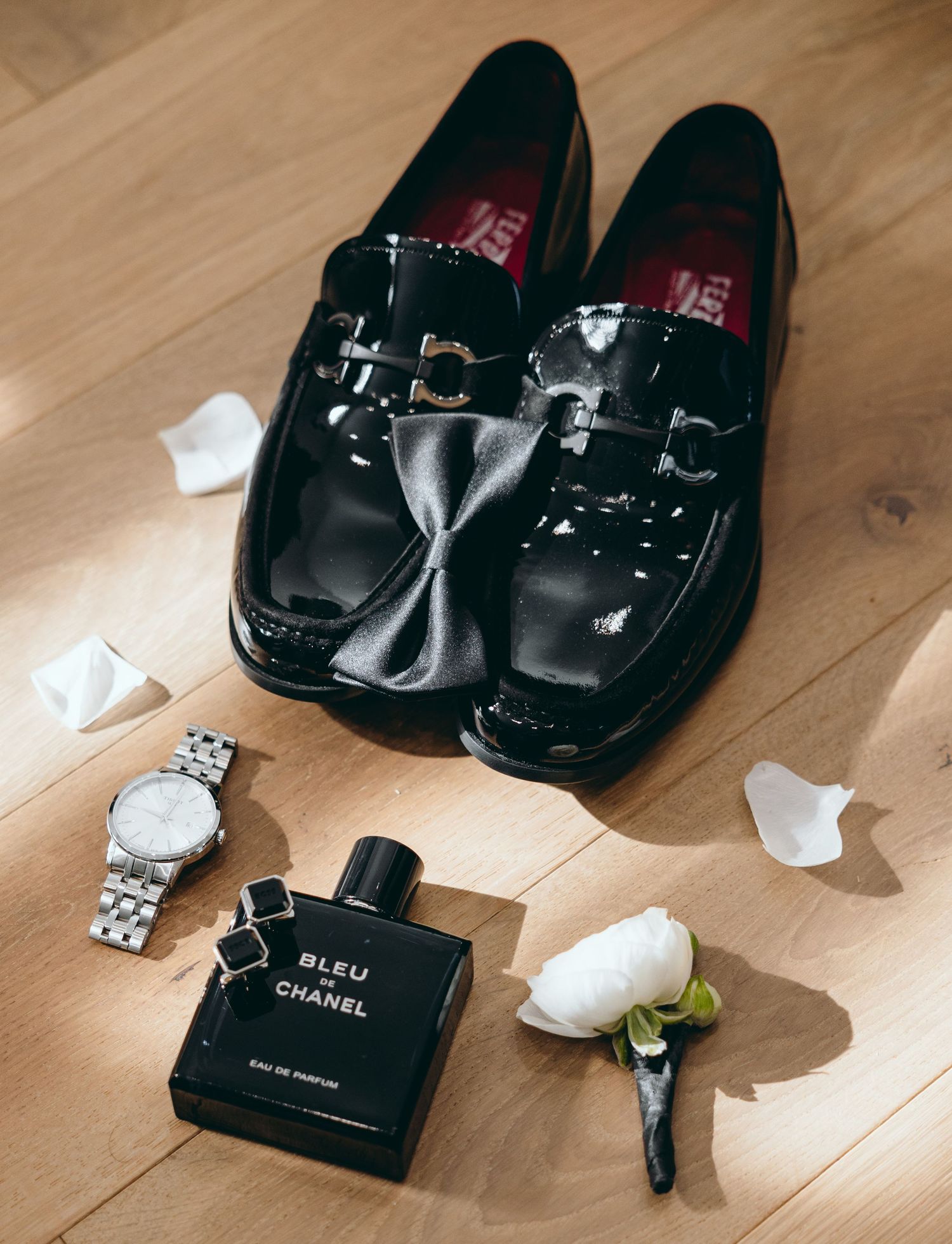 Wedding accessories including black patent leather shoes, watch, Chanel perfume and white boutonniere on wooden floor.