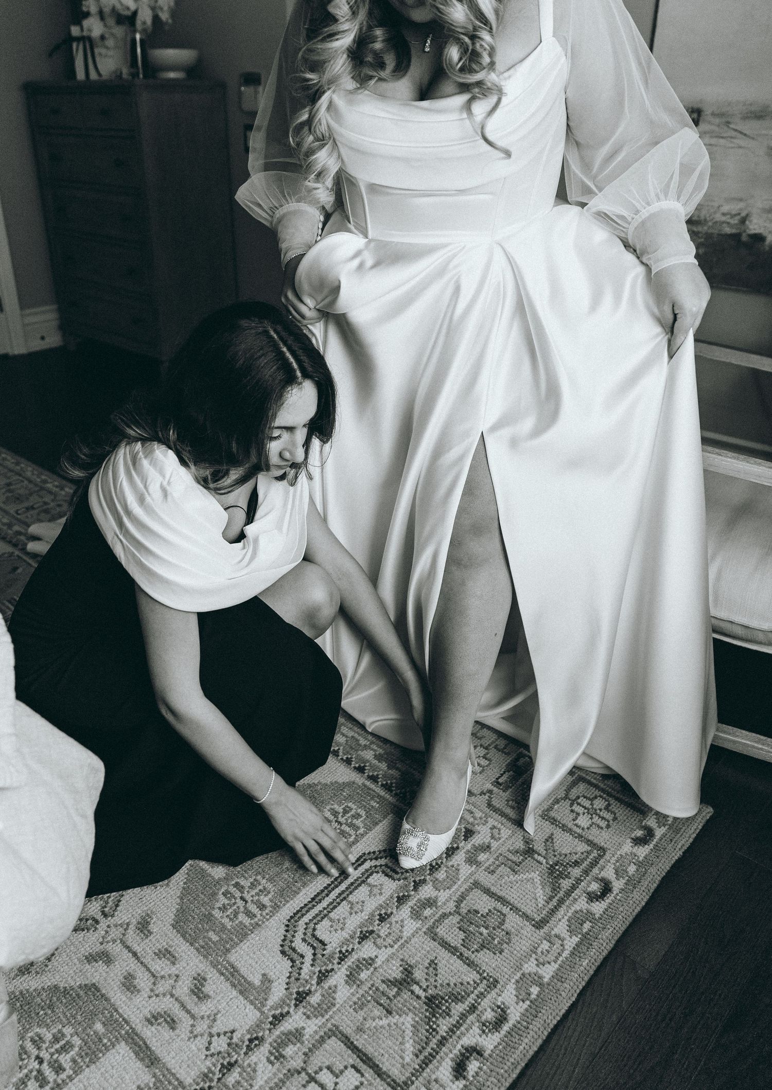 Close-up details of a white wedding dress and black bridesmaid dresses on a patterned rug.