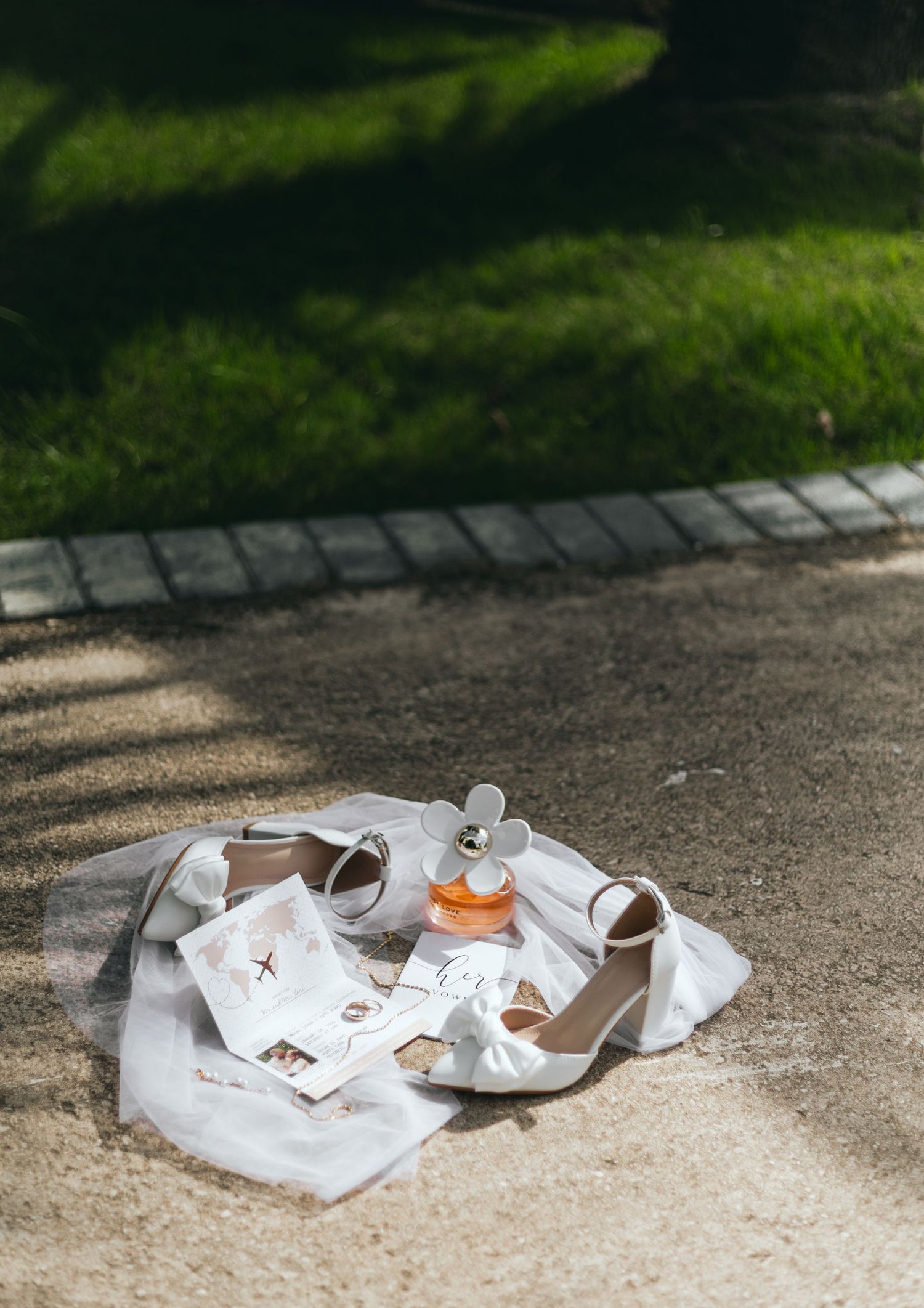 Wedding accessories and white shoes arranged on a concrete path in dappled sunlight.