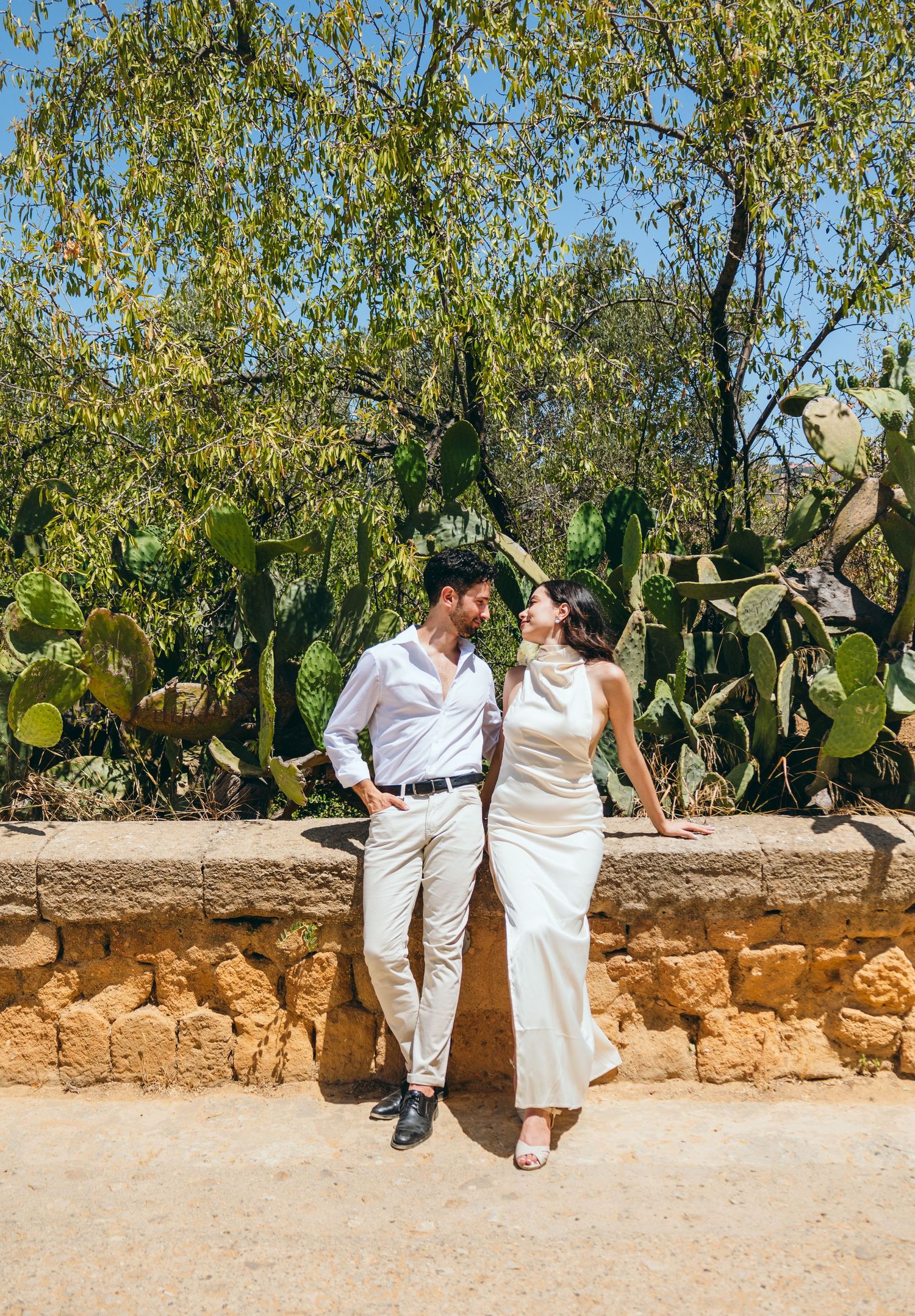 A couple in white attire sits on a stone wall surrounded by lush greenery and cacti.