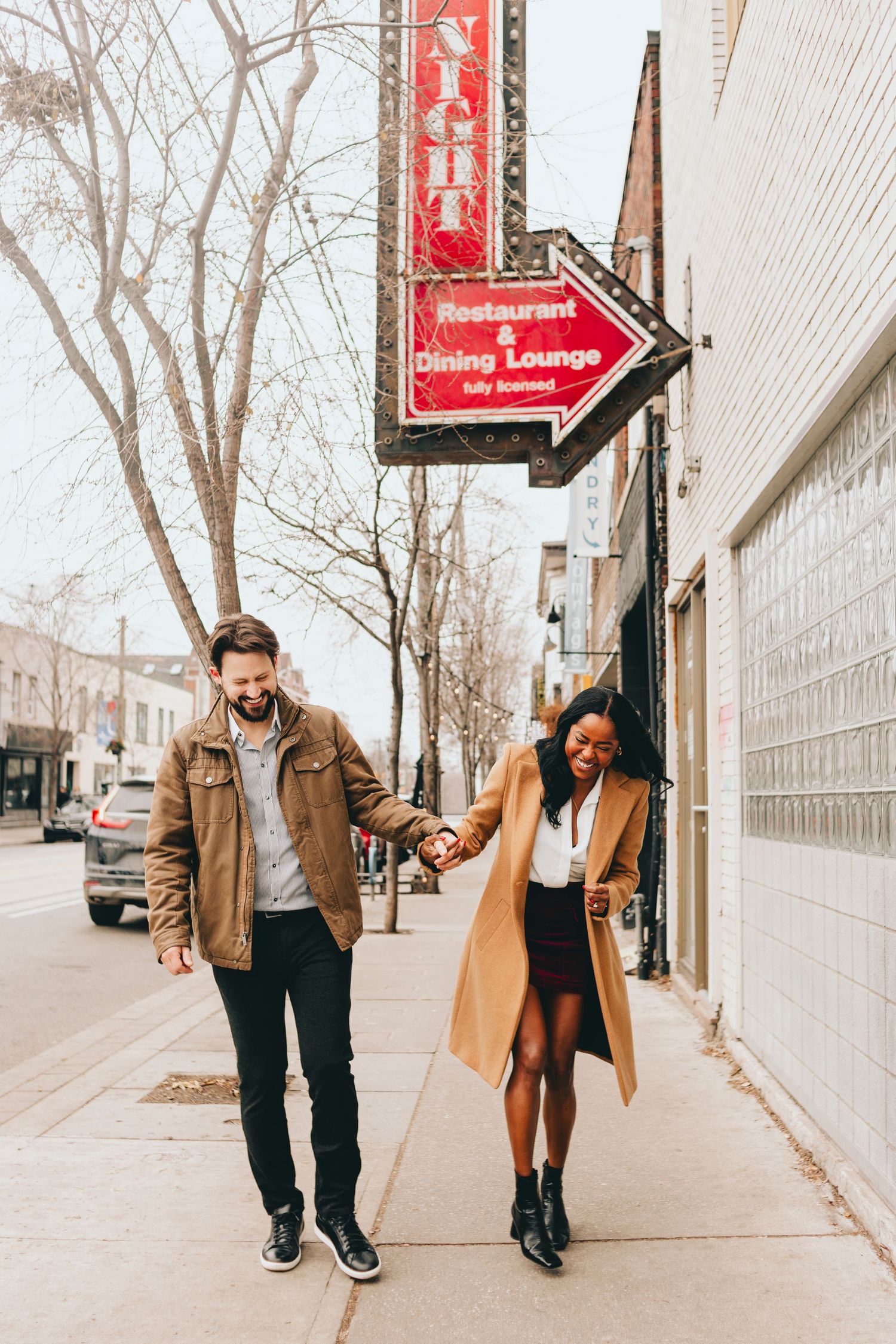 Couple walking playfully on urban sidewalk with red signage in background.