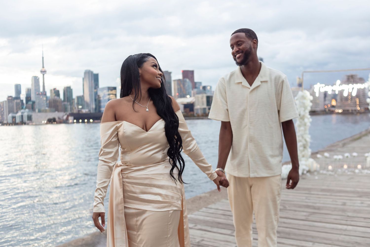 A couple in cream-colored outfits walks along a waterfront with city skyline in background.