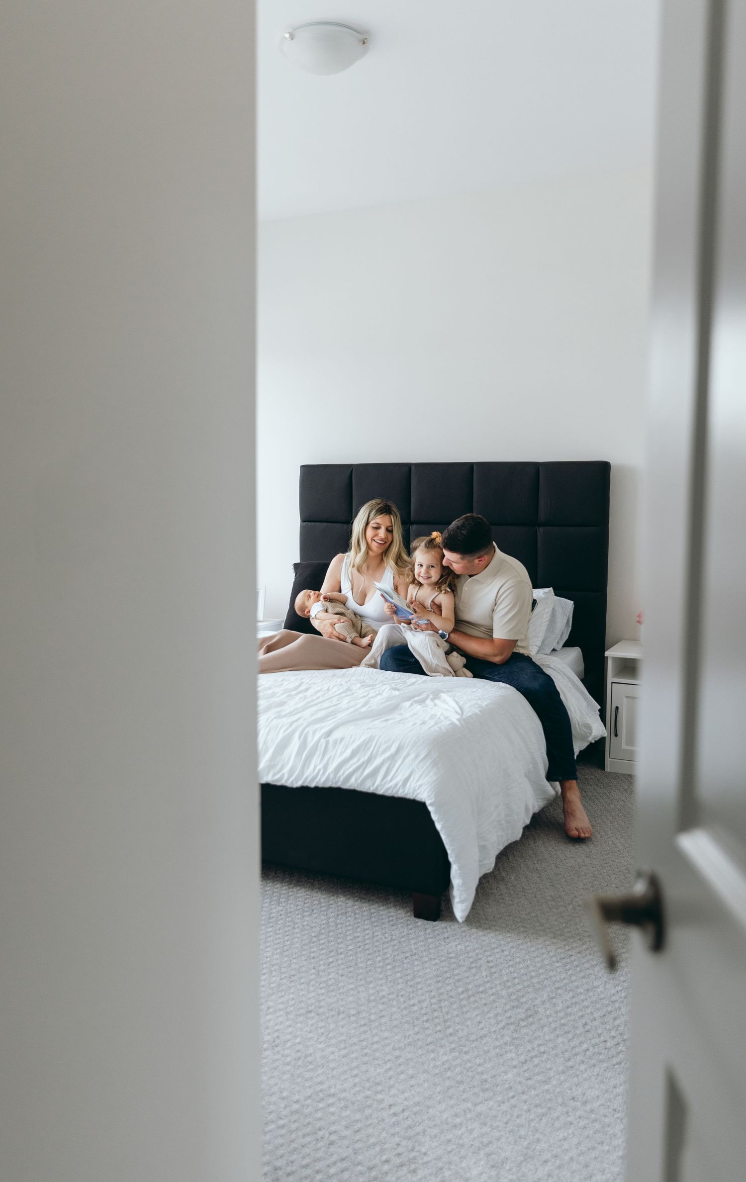 Candid family moment viewed through doorway showing bed with black headboard and white linens.