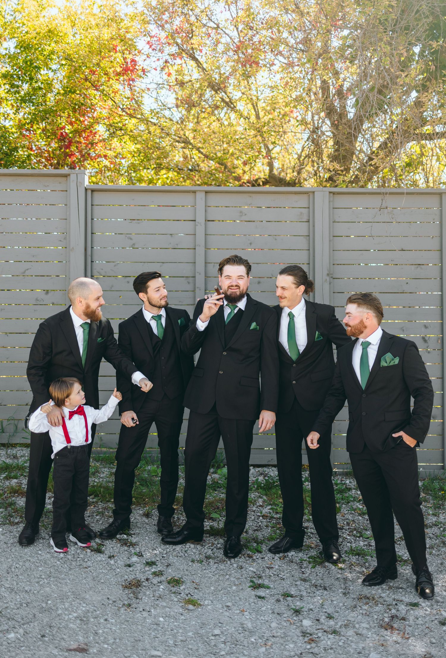 Wedding party in black suits with green ties posing casually against a grey fence.