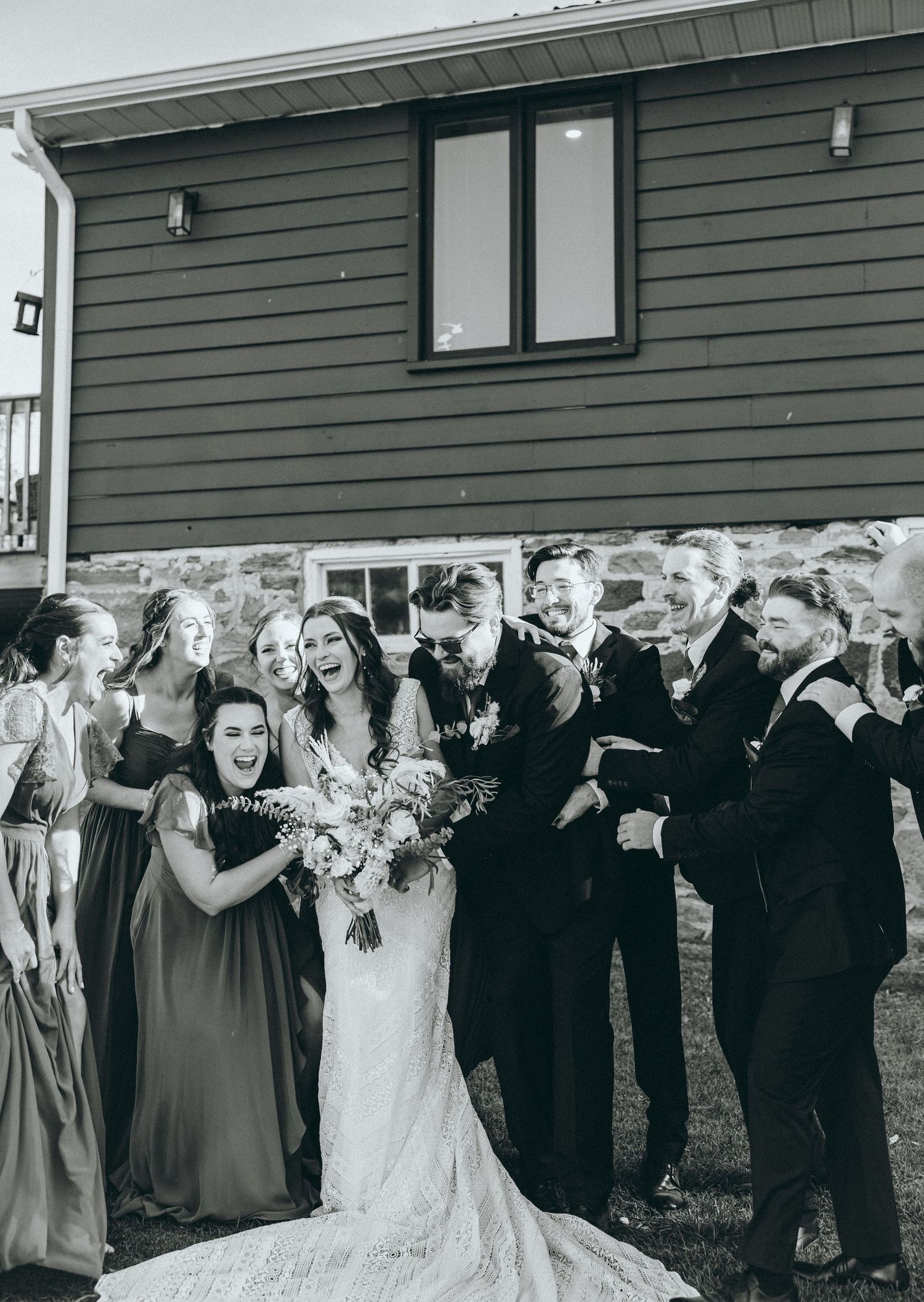 Wedding party members strike fun poses in a line against grey building exterior.