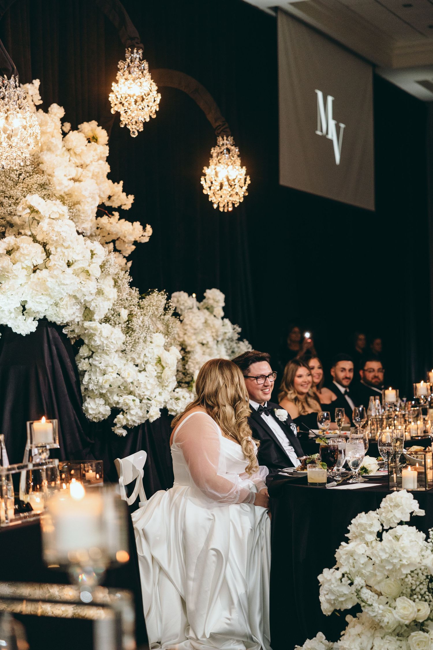 Elegant wedding reception setup with crystal chandeliers and white floral arrangements against a dark backdrop.
