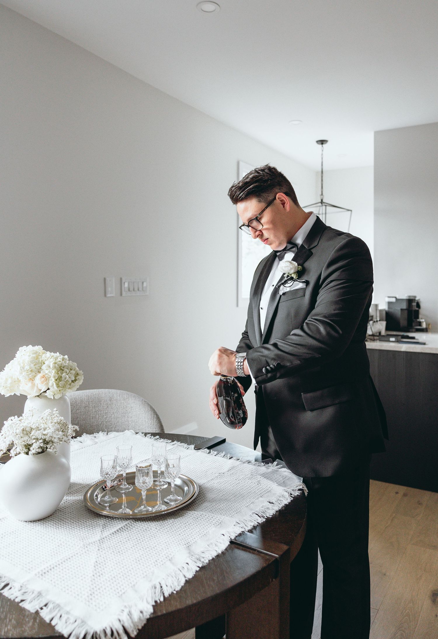 A person in a black suit adjusts their tie while standing in a minimalist white room.