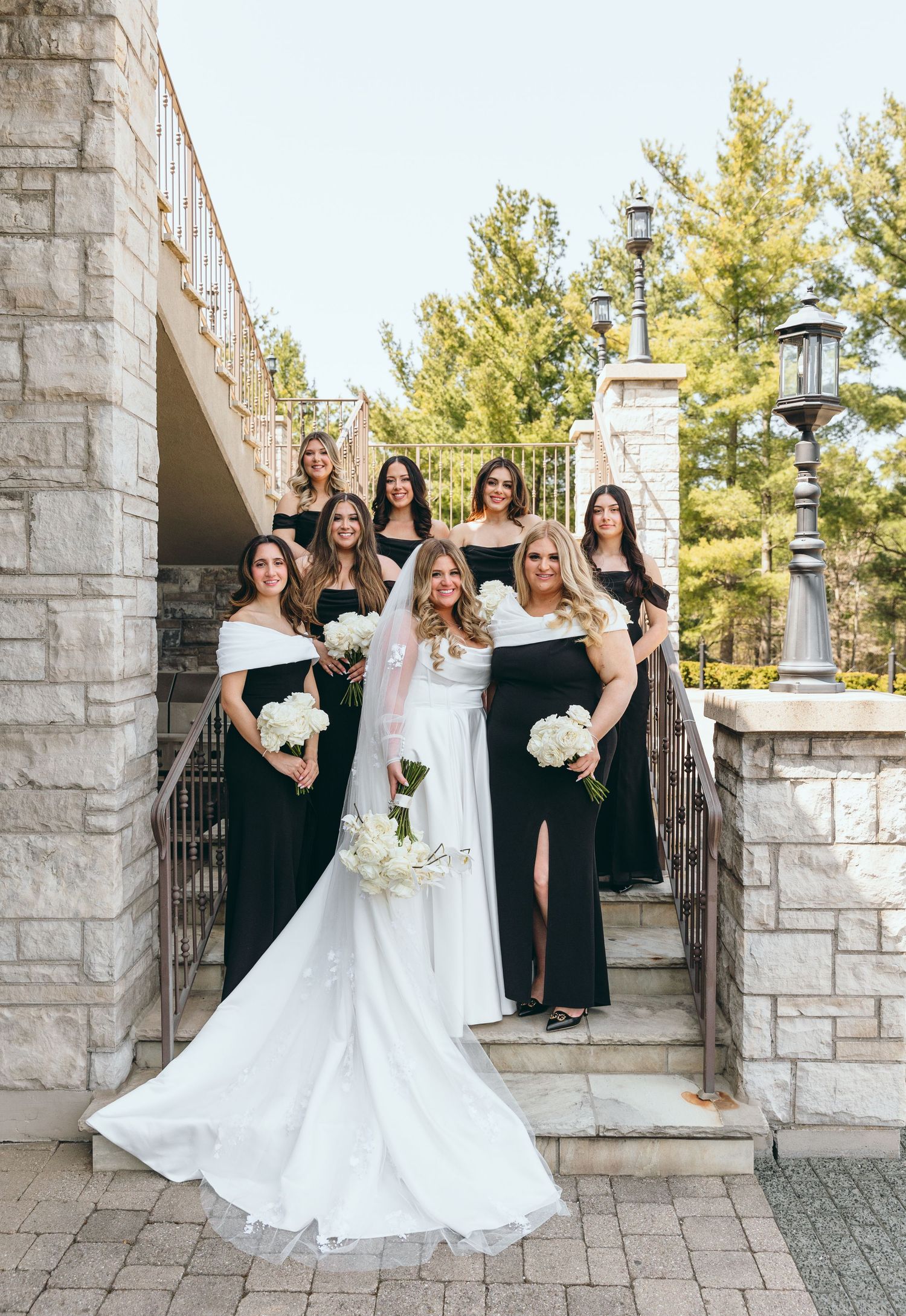 Bridal party in black dresses celebrates with bride in flowing white gown on stone steps.