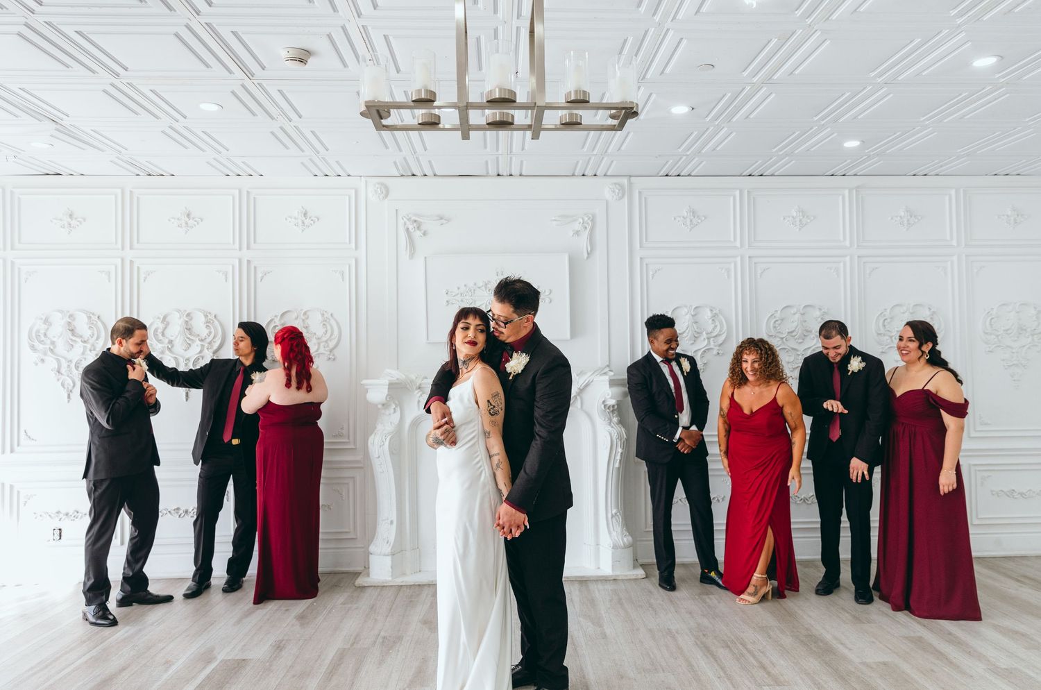 Wedding party poses playfully in a bright white room with burgundy and black attire.