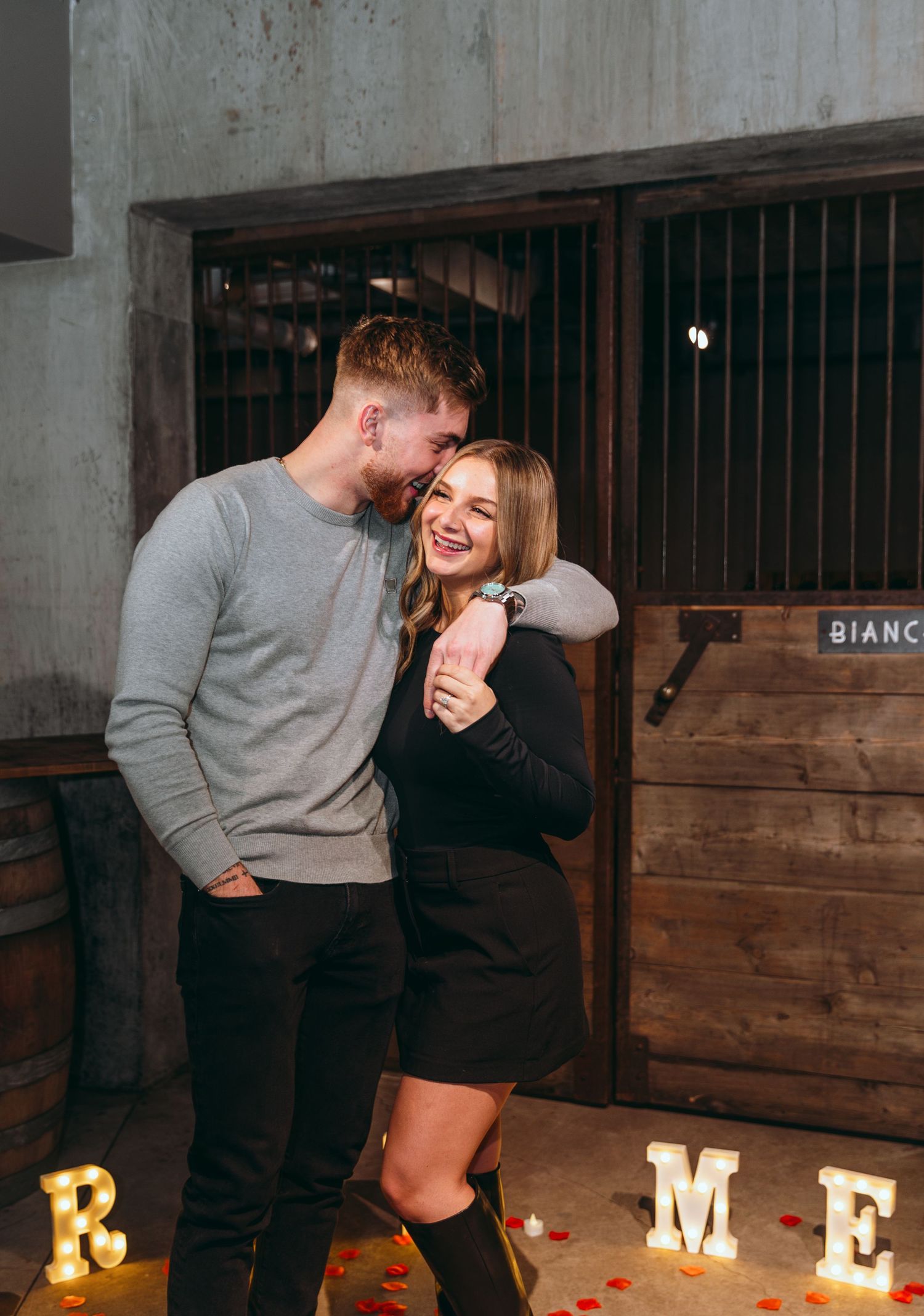 A romantic proposal scene in a rustic indoor setting with glowing letters and rose petals on the floor.