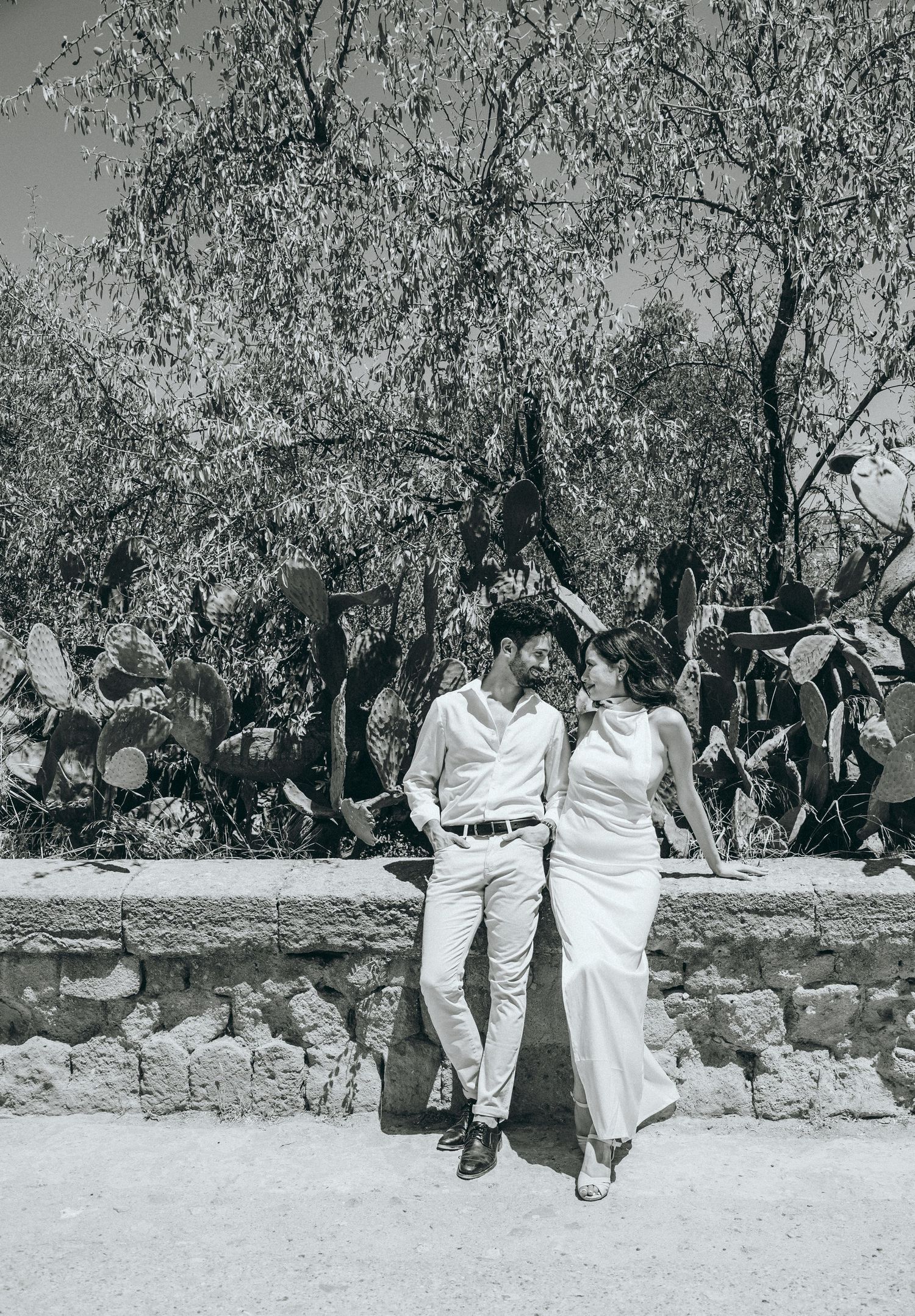 Couple walking together along stone wall beneath leafy trees in black and white.