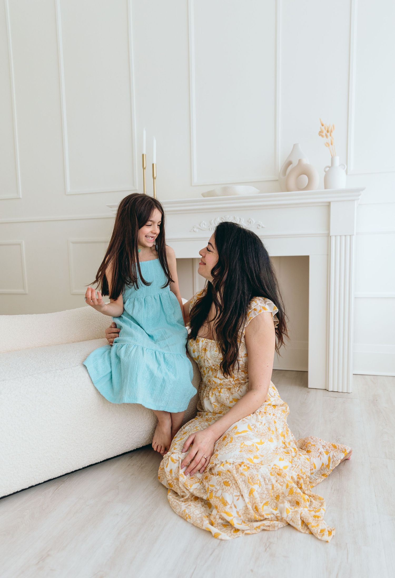 Two women in floral and turquoise dresses laughing together on light wooden floor near white fireplace.