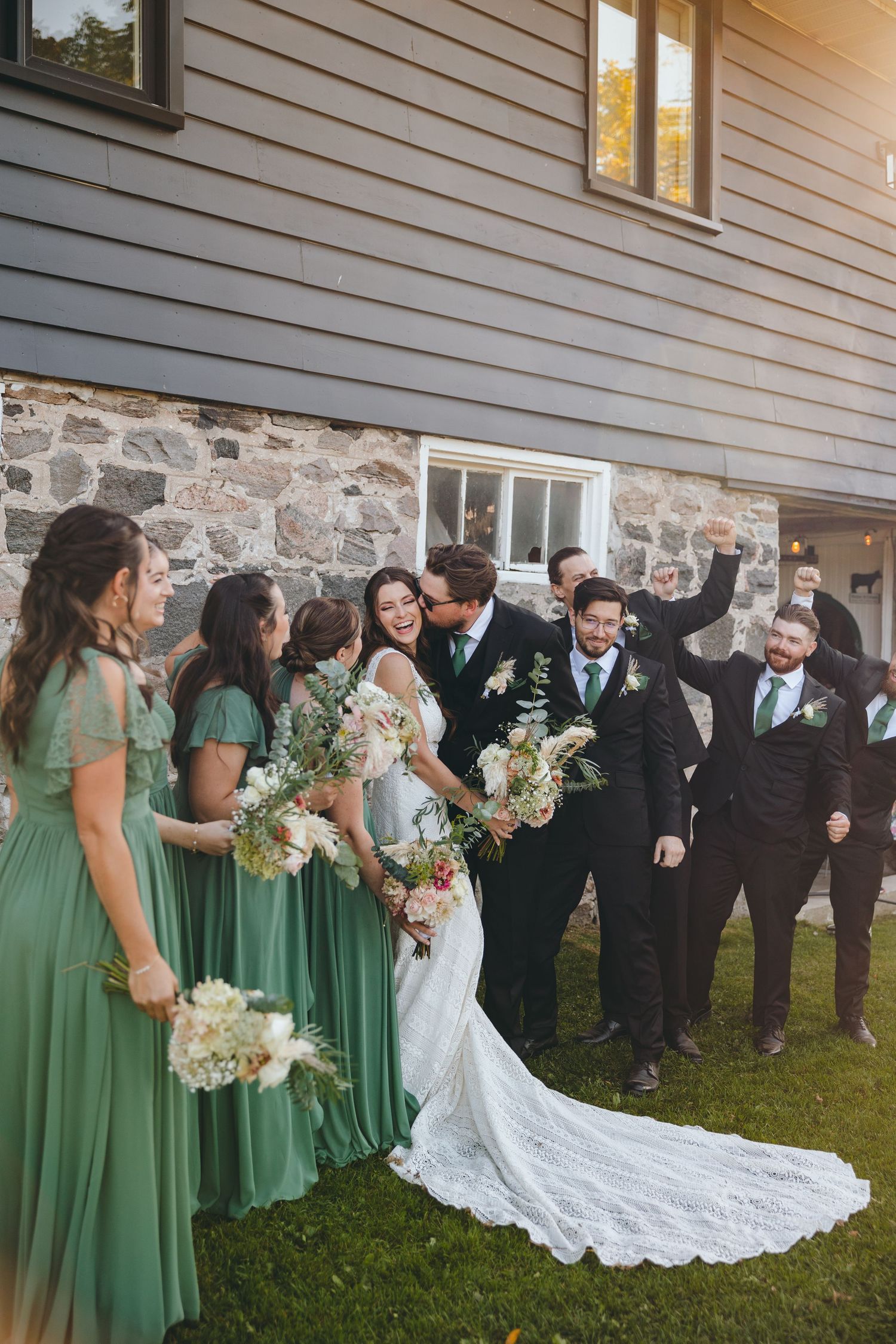 Wedding party in emerald green dresses and black suits posed against stone building exterior.
