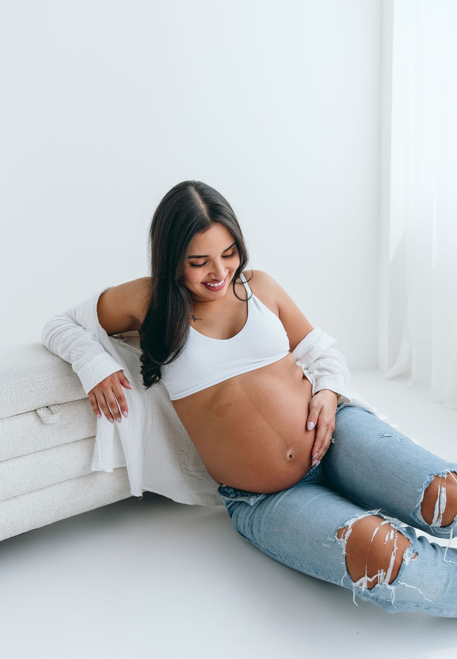 Expecting mother in ripped jeans poses against white backdrop during studio photoshoot.