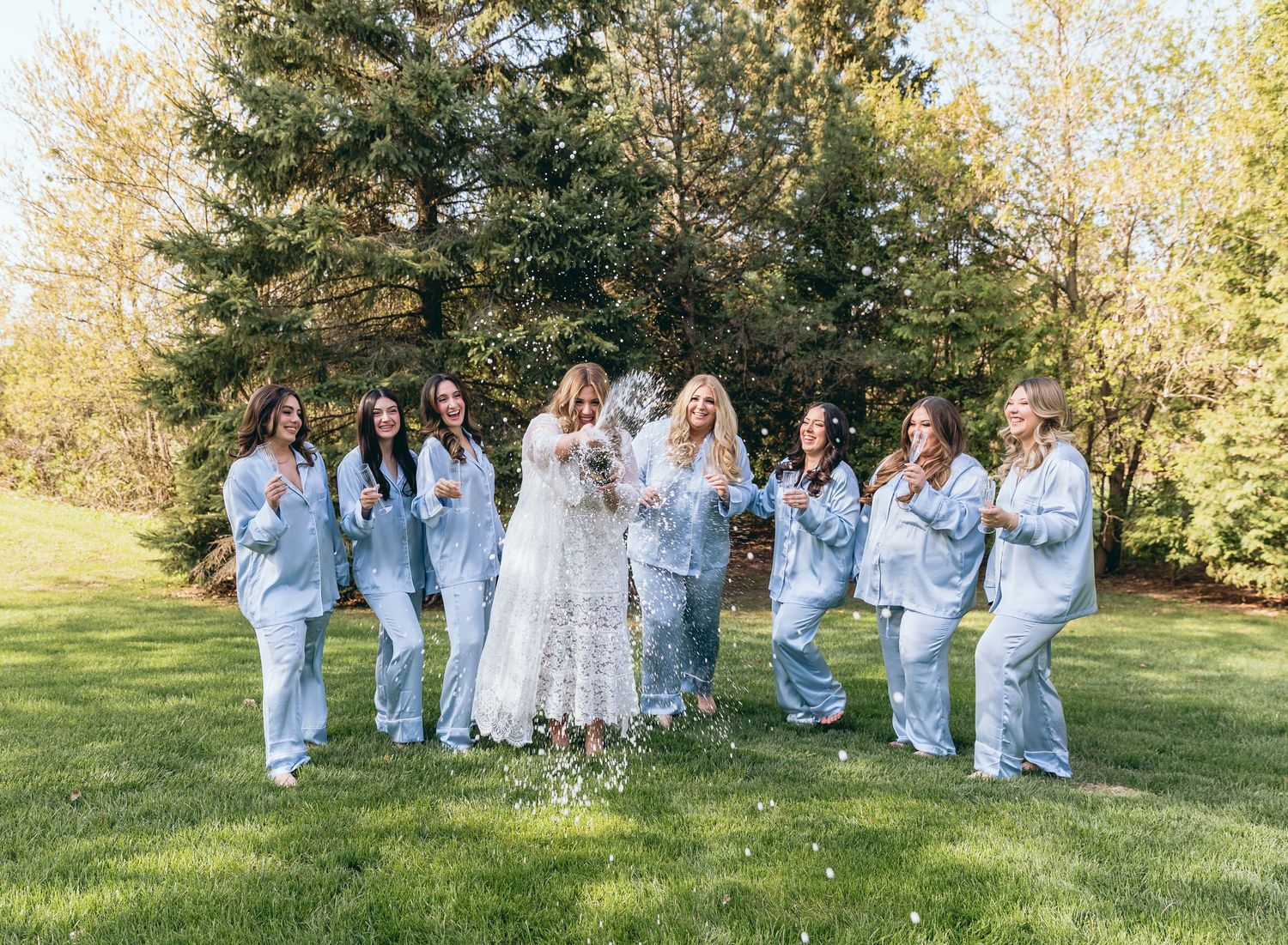 Bridal party in white dresses and denim jackets dancing together on grass in sunlight.