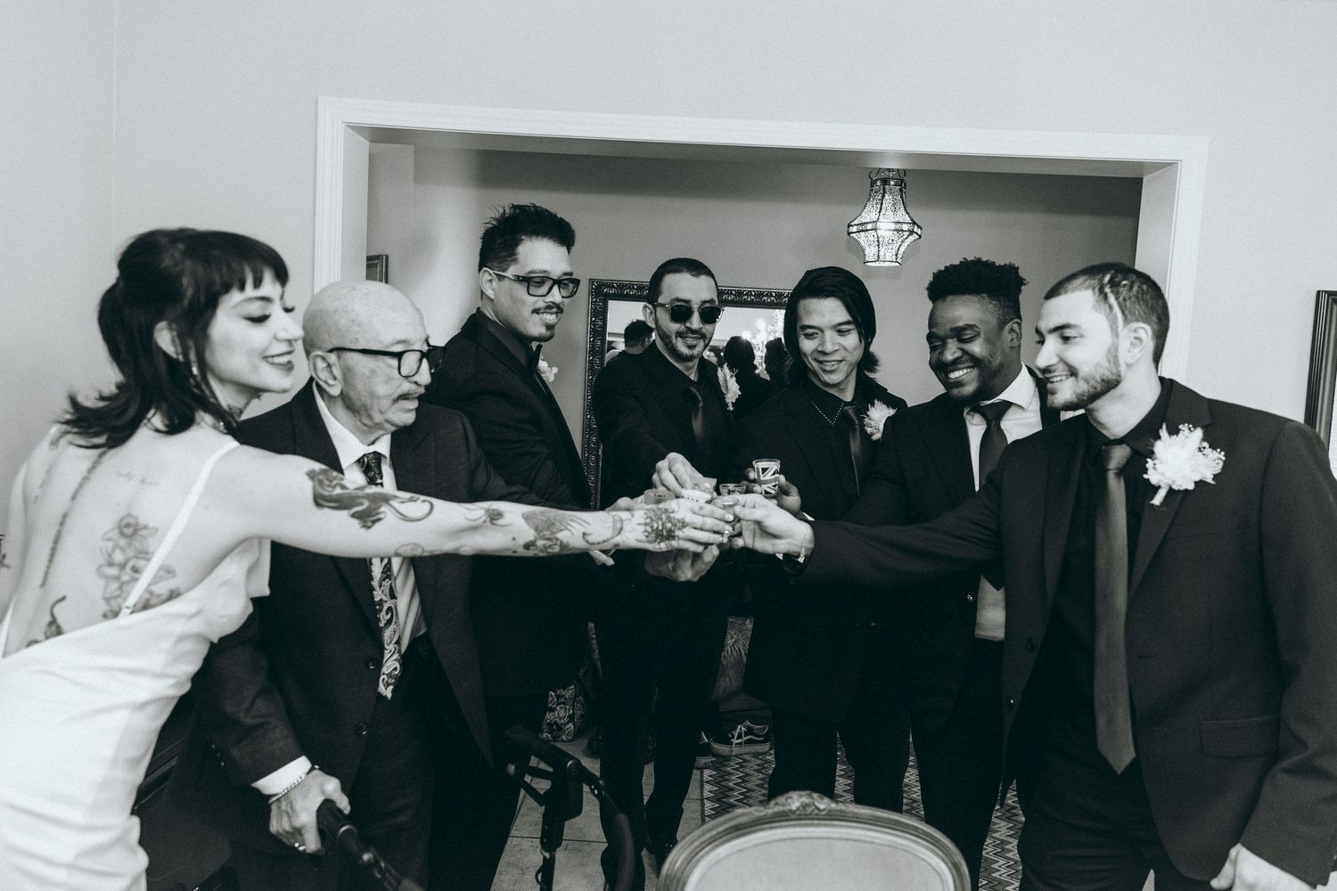 Wedding party members in black suits and burgundy dresses pose together in a white room.