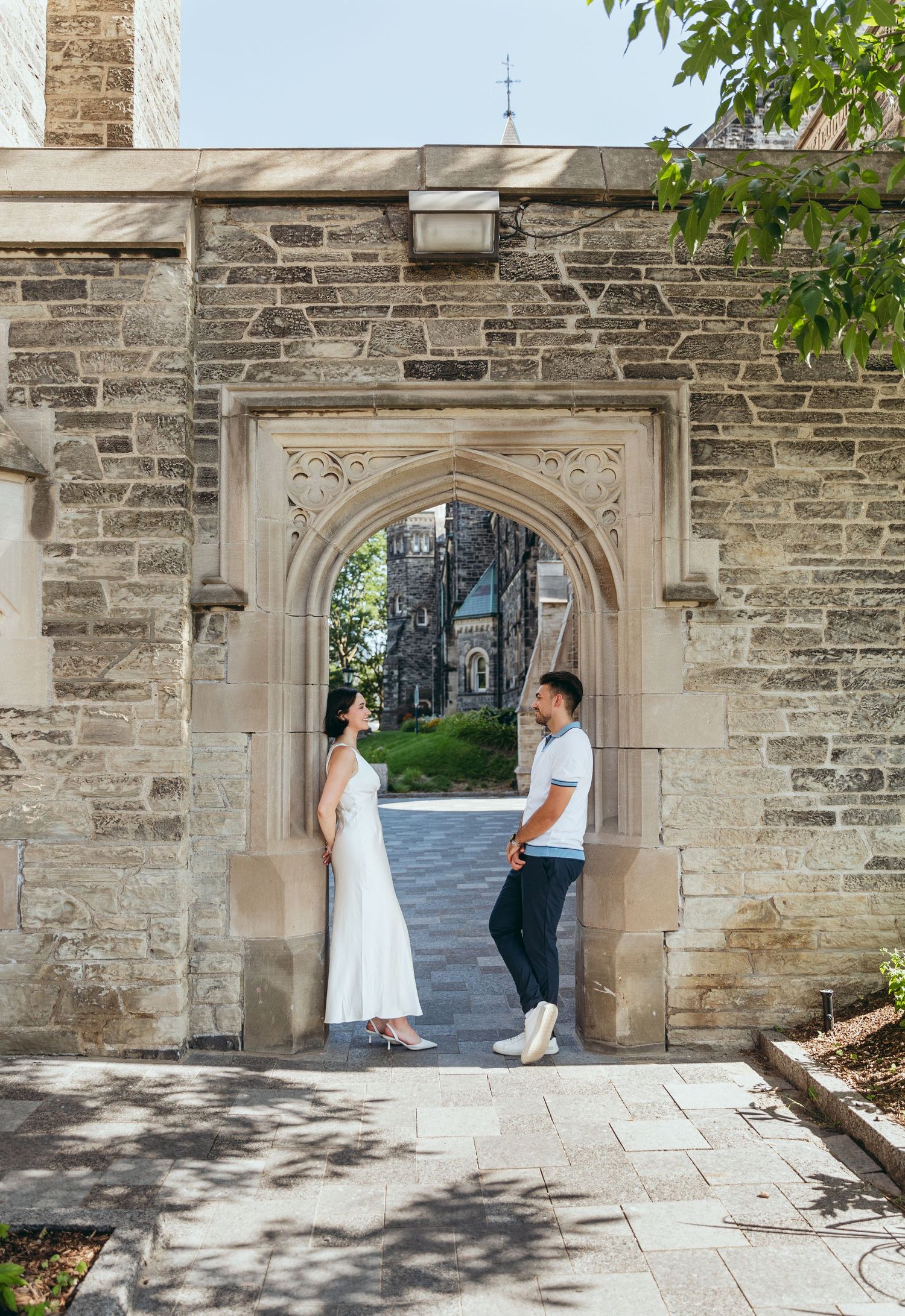 A couple stands facing each other in a gothic stone doorway on a bright summer day.