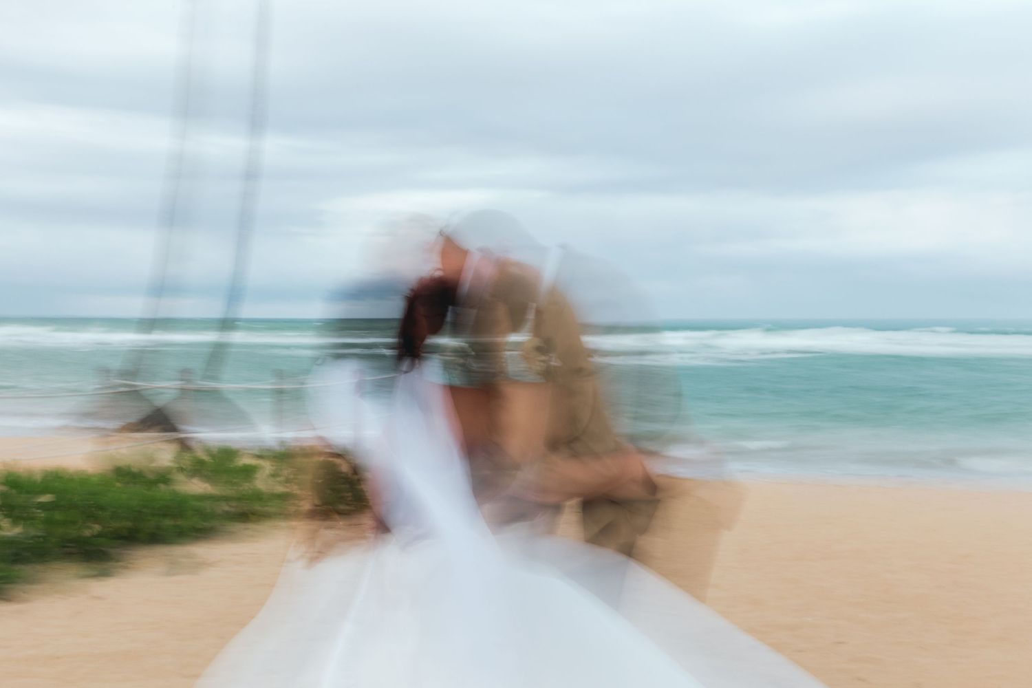 A couple shares a romantic dip kiss on a tropical beach with palm trees and turquoise waters in the background.