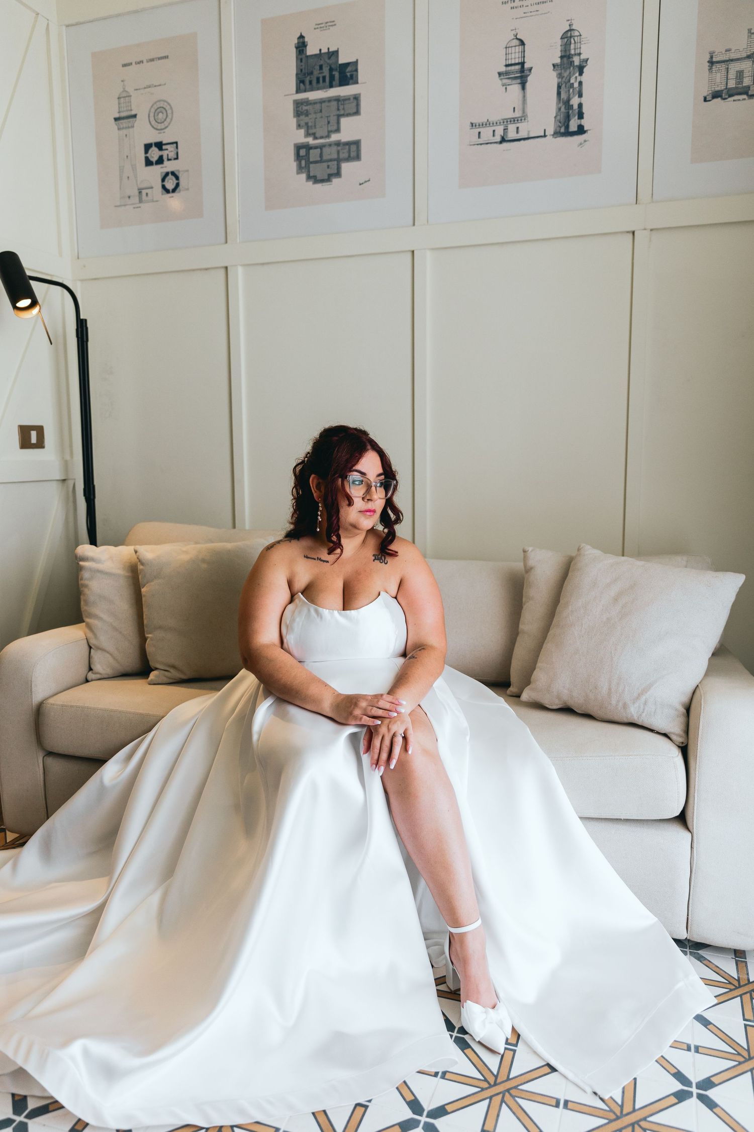 Person in white wedding dress poses on beige couch against wall with framed architectural prints.