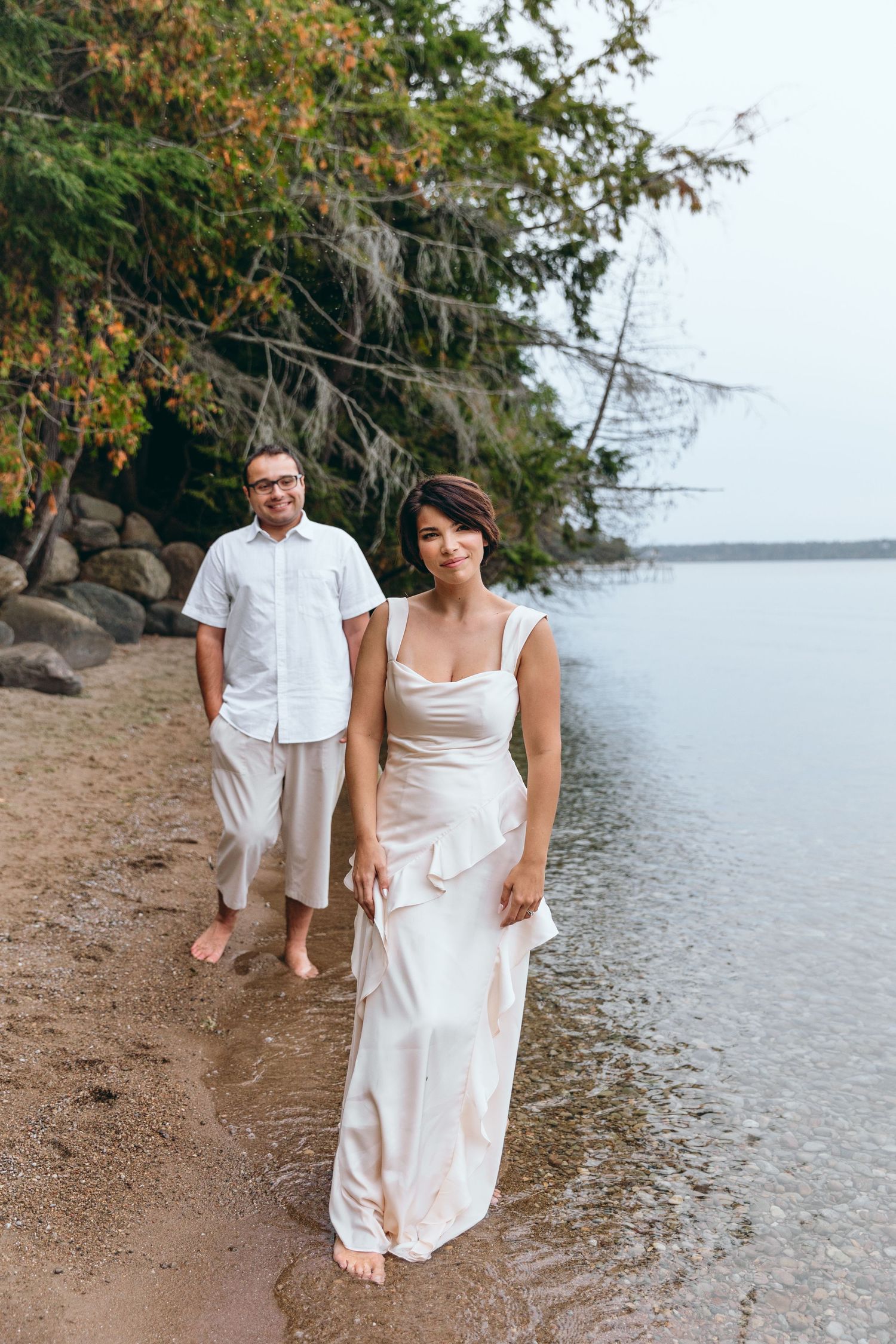 Couple in white outfits walking along shoreline of lake with trees in background.