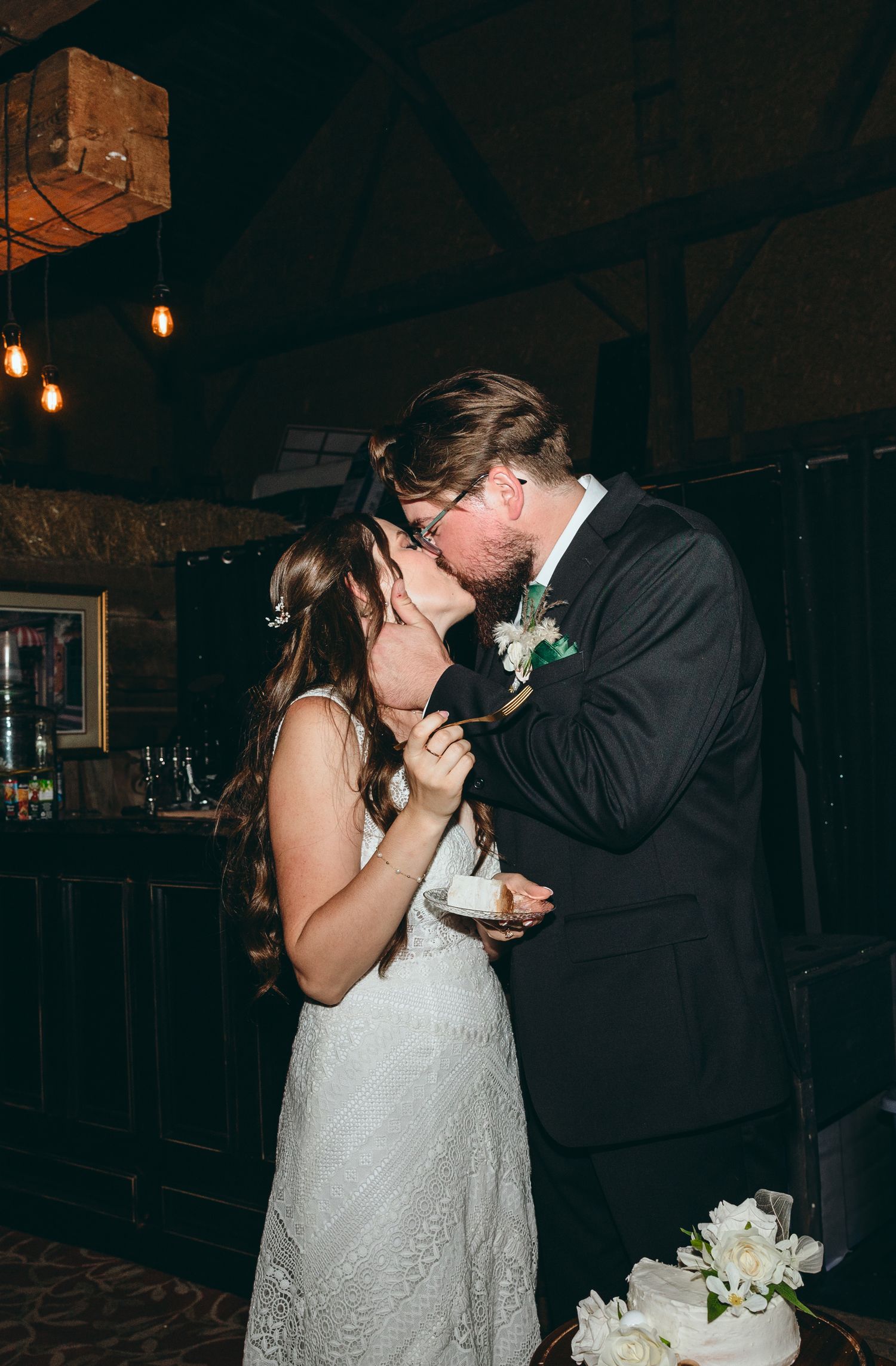 Couple sharing first bite of wedding cake in dimly lit rustic venue.