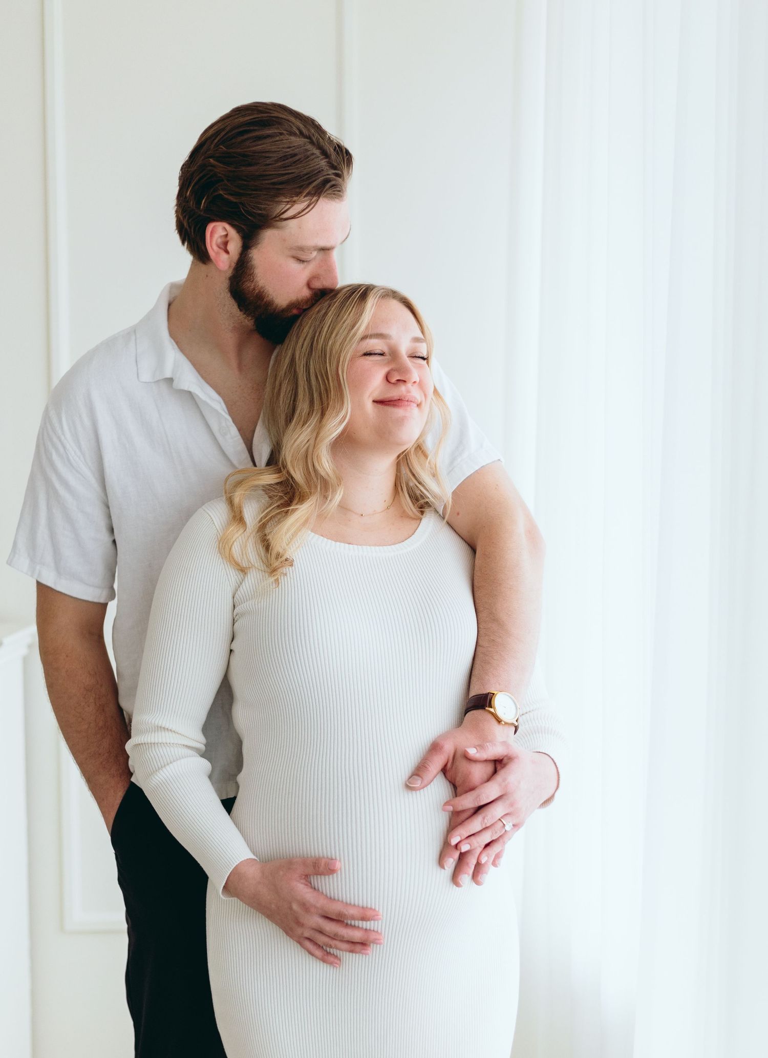 Tender moment between expecting couple in white clothing by window.