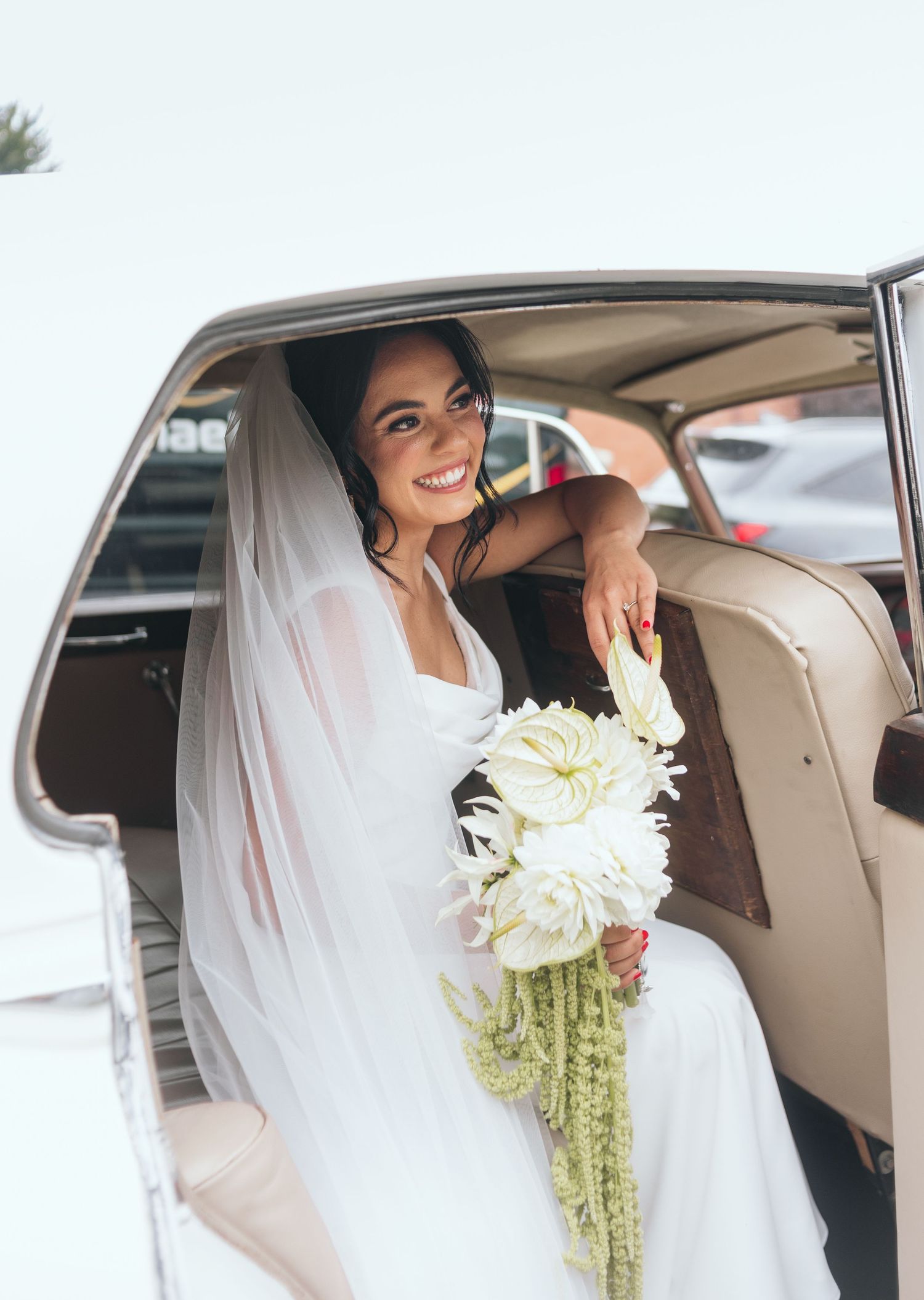 A bride in a white dress and veil sits in a luxury car holding a white bouquet.
