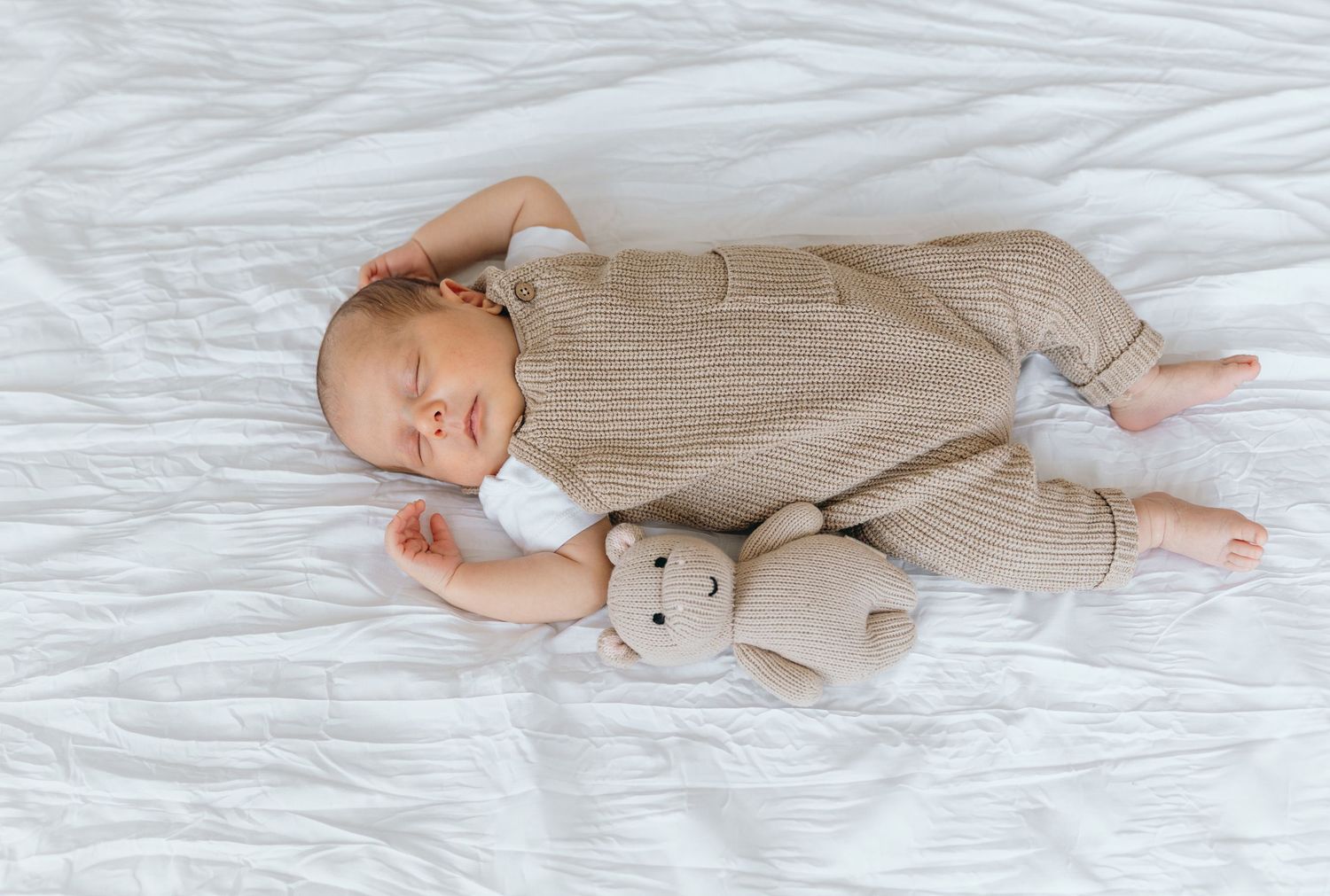 Sleeping infant in beige knit outfit lying on white bedding with plush teddy bear companion.