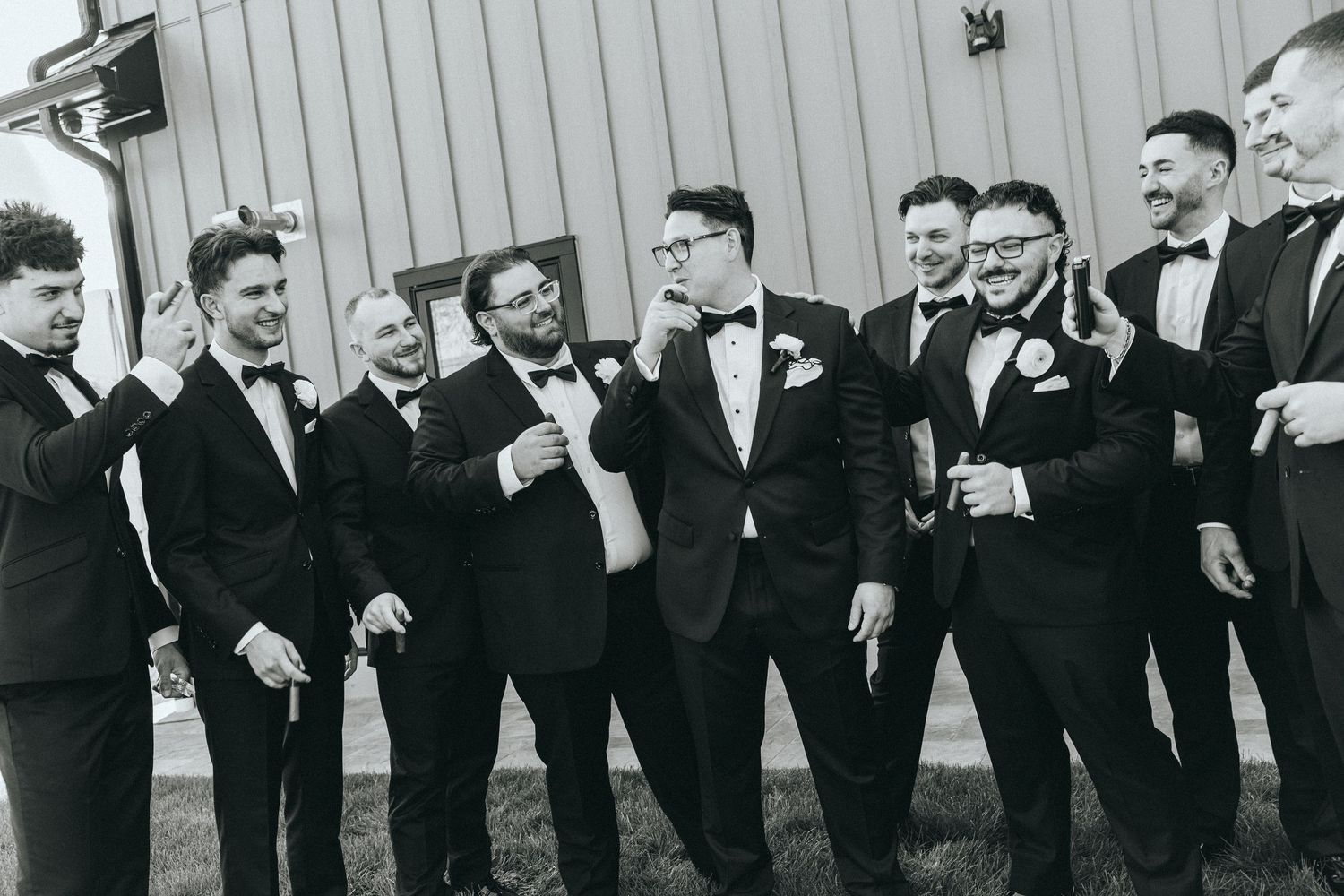 Line of groomsmen in black suits posing against white building exterior.