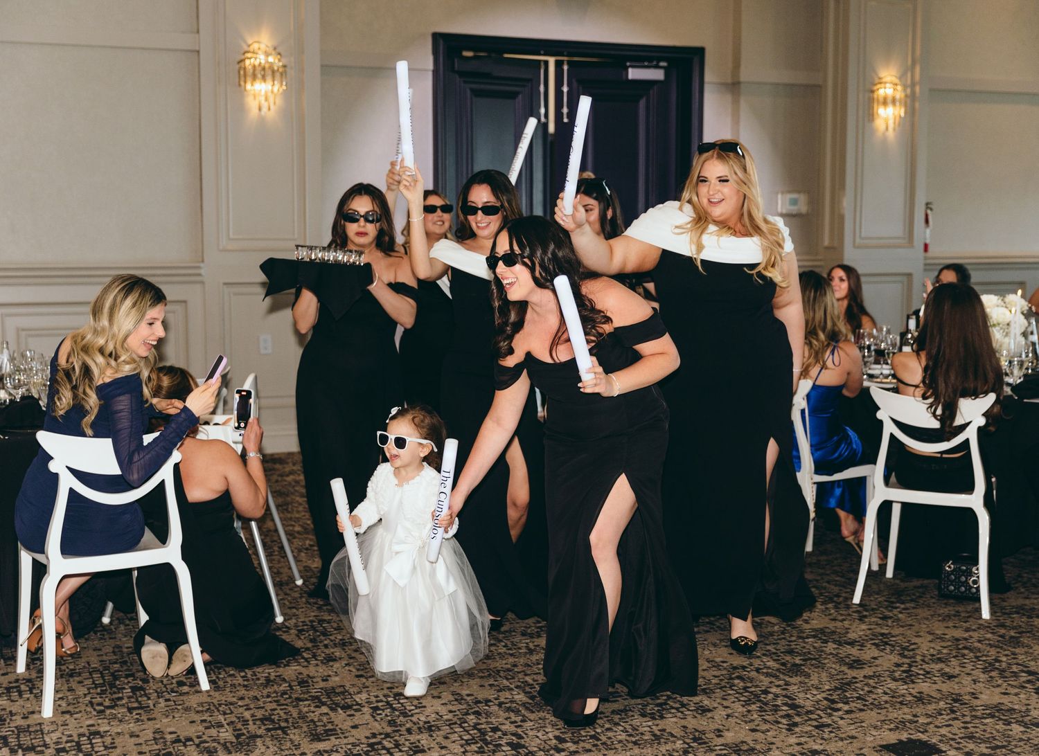 Wedding party in black dresses dancing with flower girl in white dress at reception.