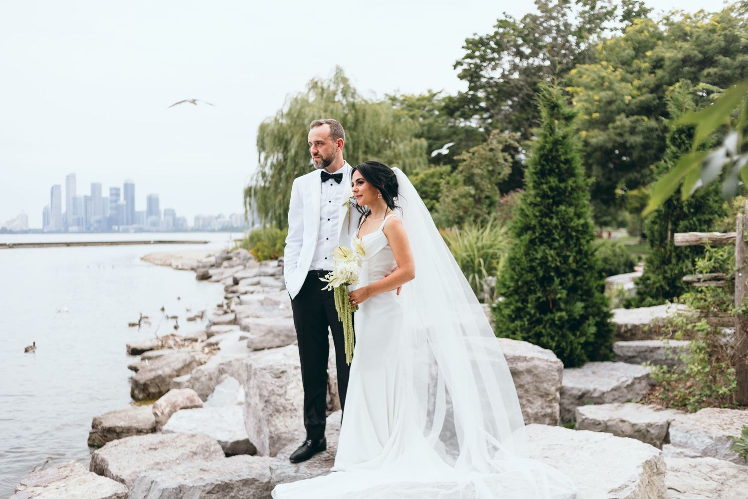 A bride and groom pose together on a rocky shoreline with a city skyline visible across the water.
