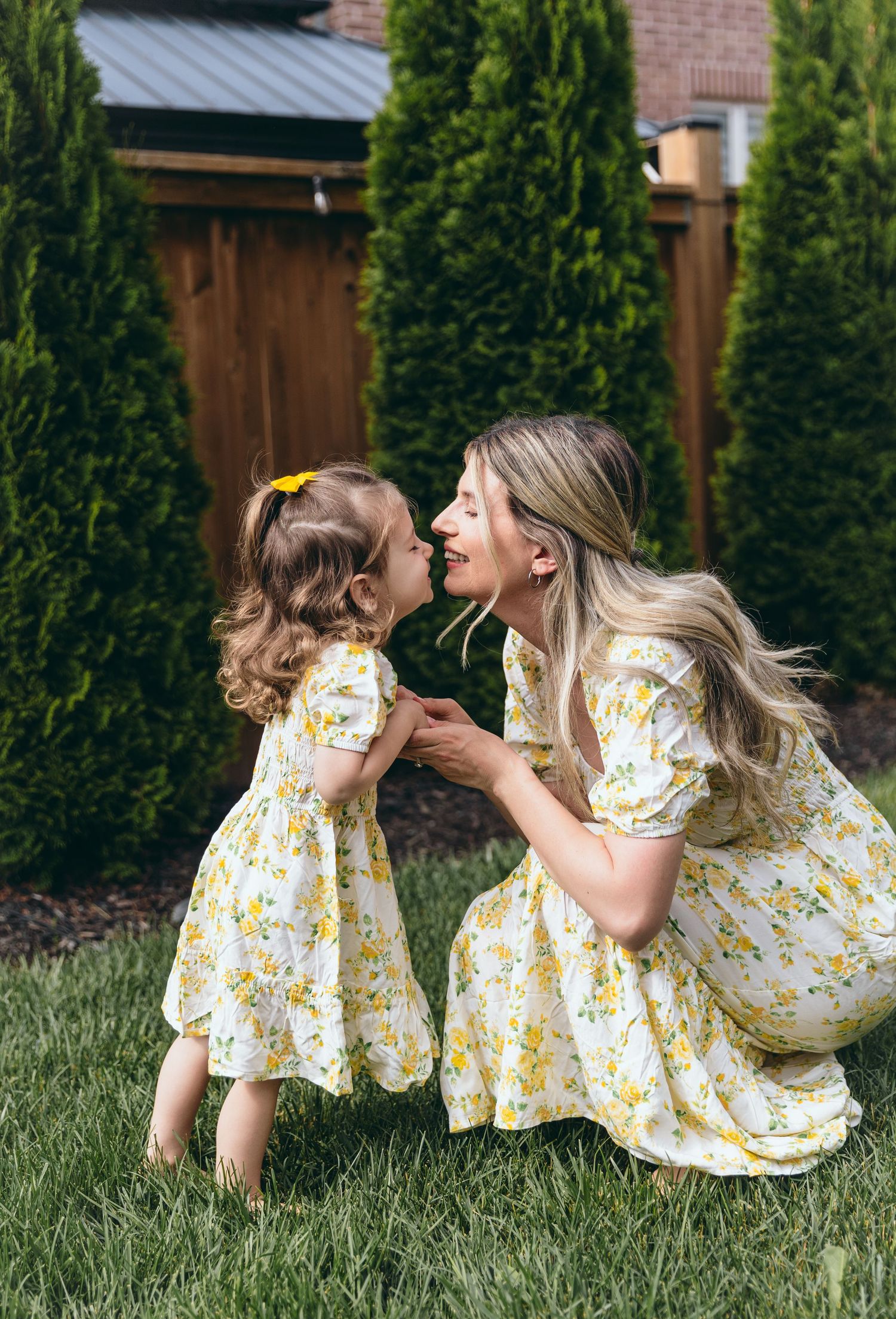 Mother and daughter in matching yellow floral dresses playing together on green grass.