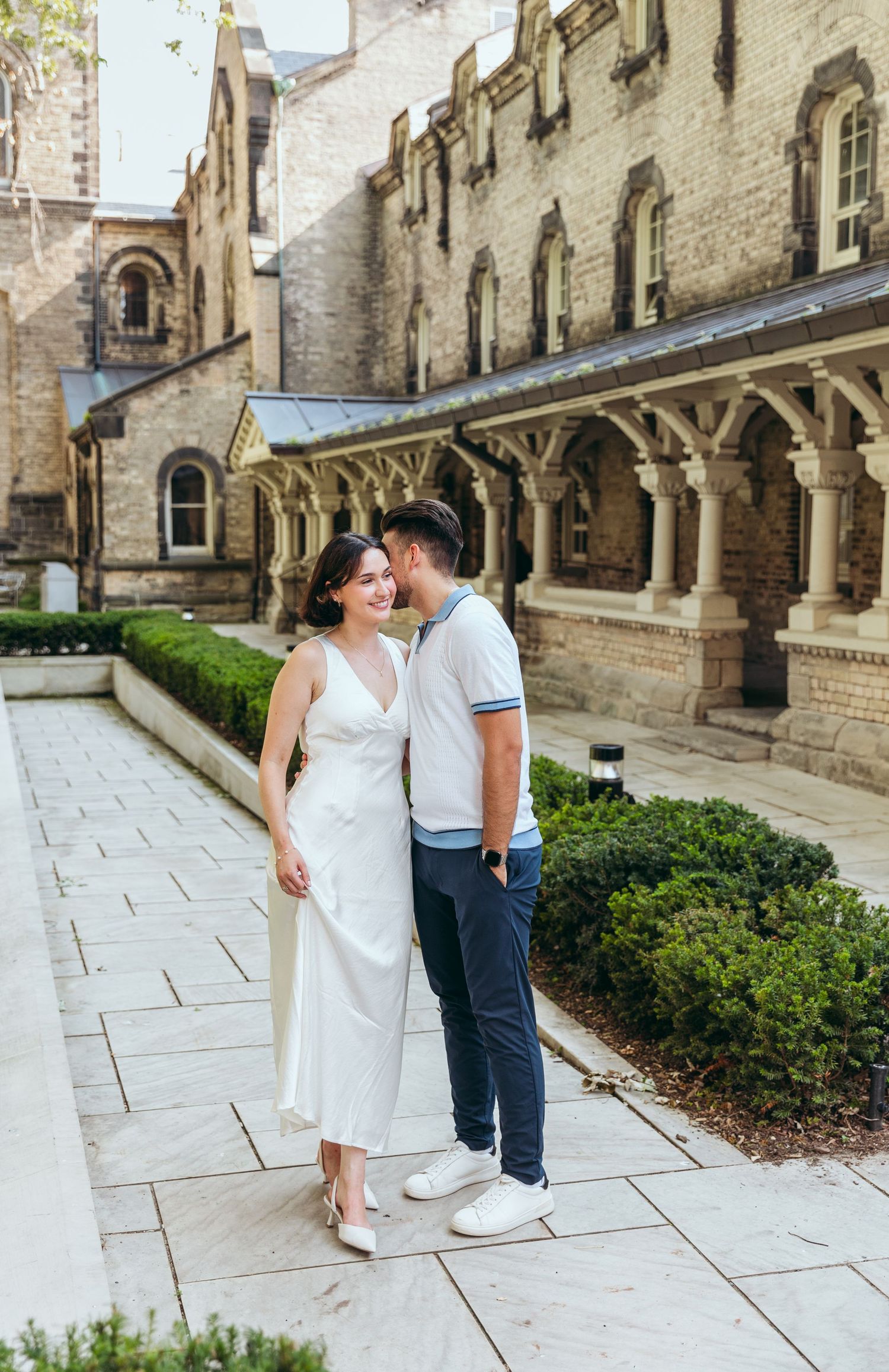 A couple stands together on a stone pathway beside Gothic architecture and well-maintained gardens.