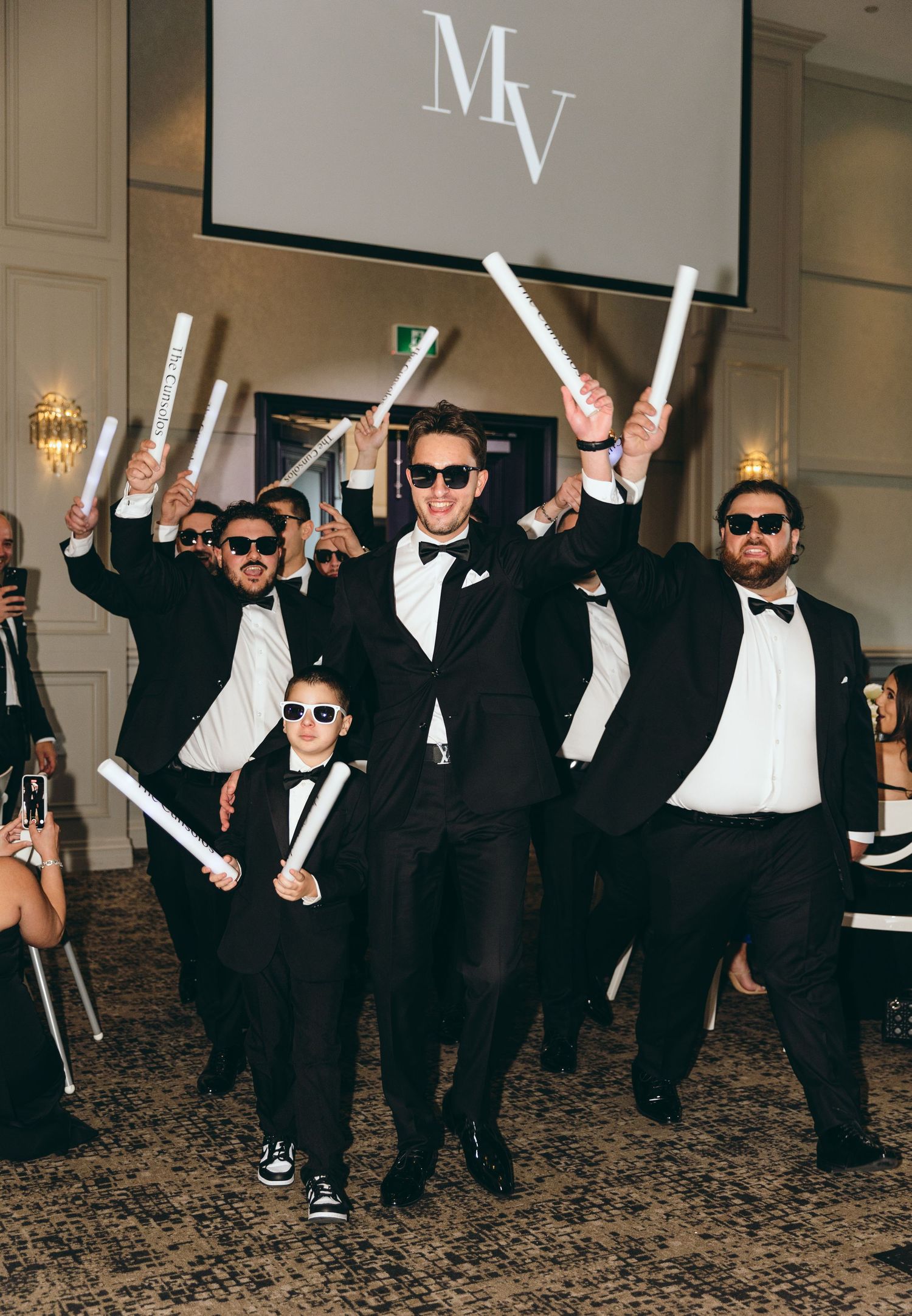 Group in formal attire and sunglasses posing with light-up batons at wedding reception.