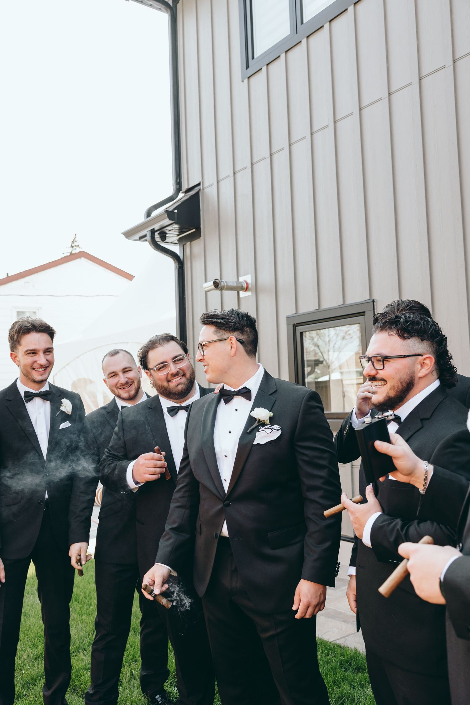 Groomsmen in black tuxedos share laughs outside a building during wedding festivities.