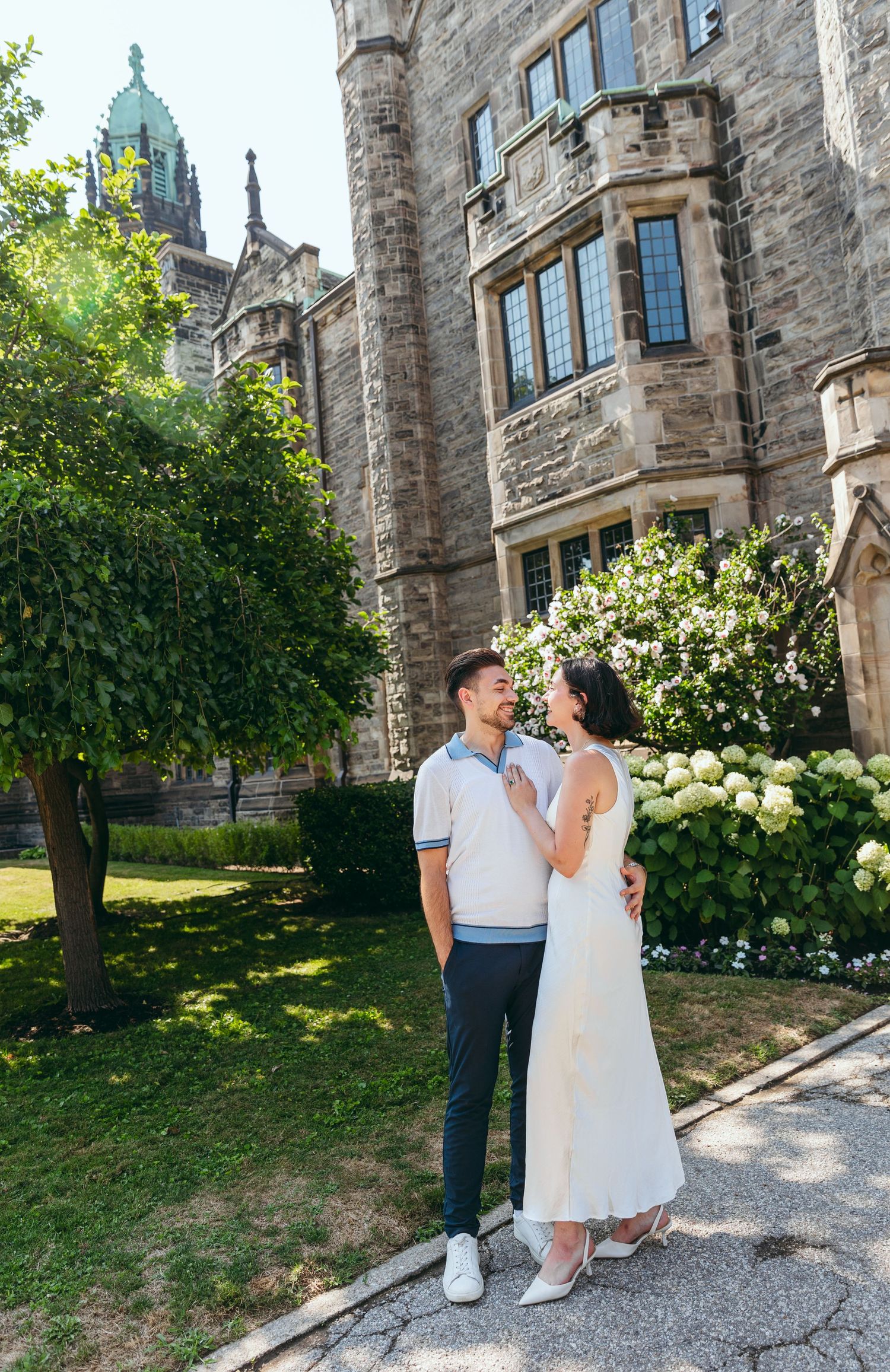 A couple poses together outside a historic stone building with blooming hydrangeas.