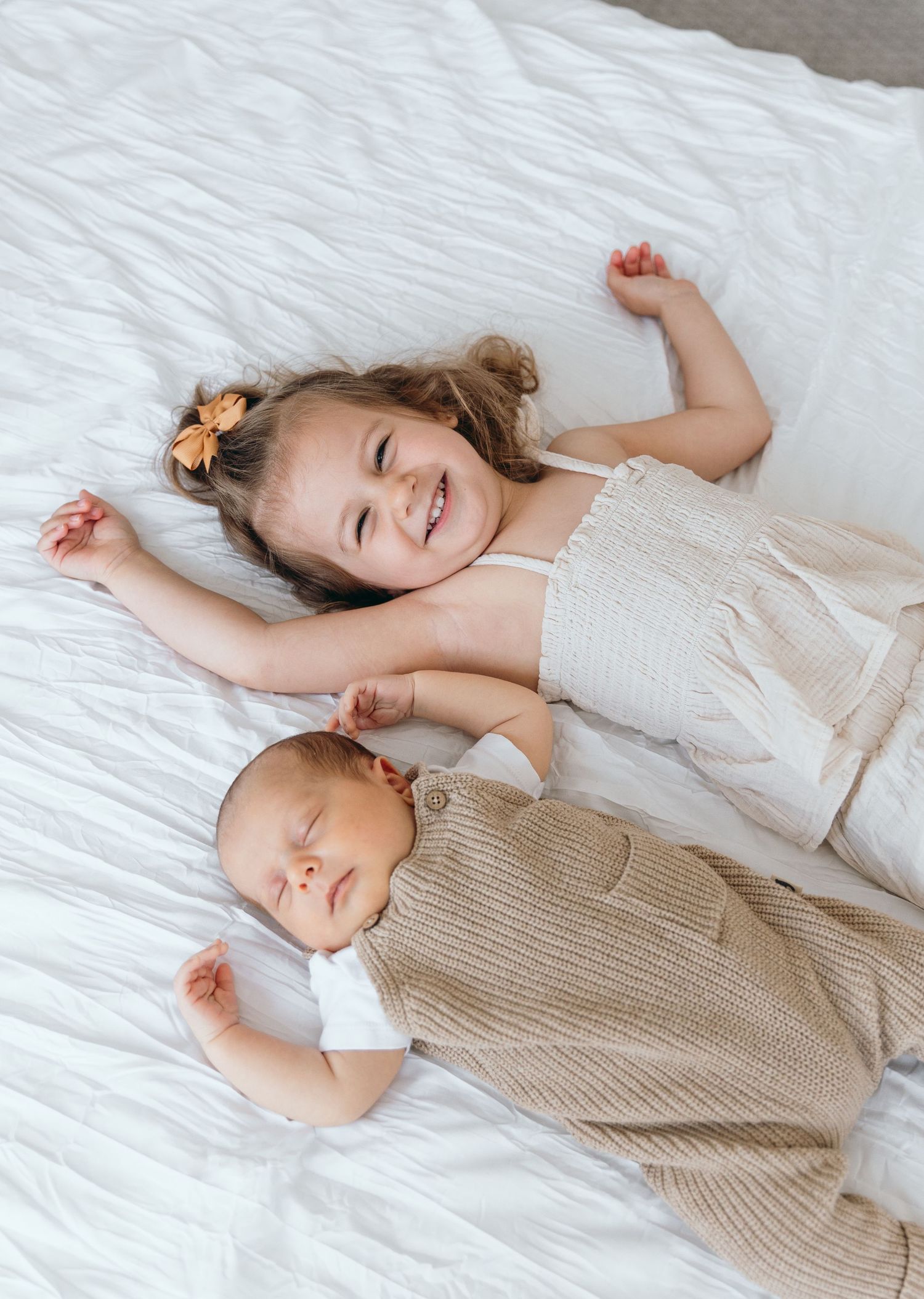 Two children lying peacefully on white bedding wearing neutral colored outfits.