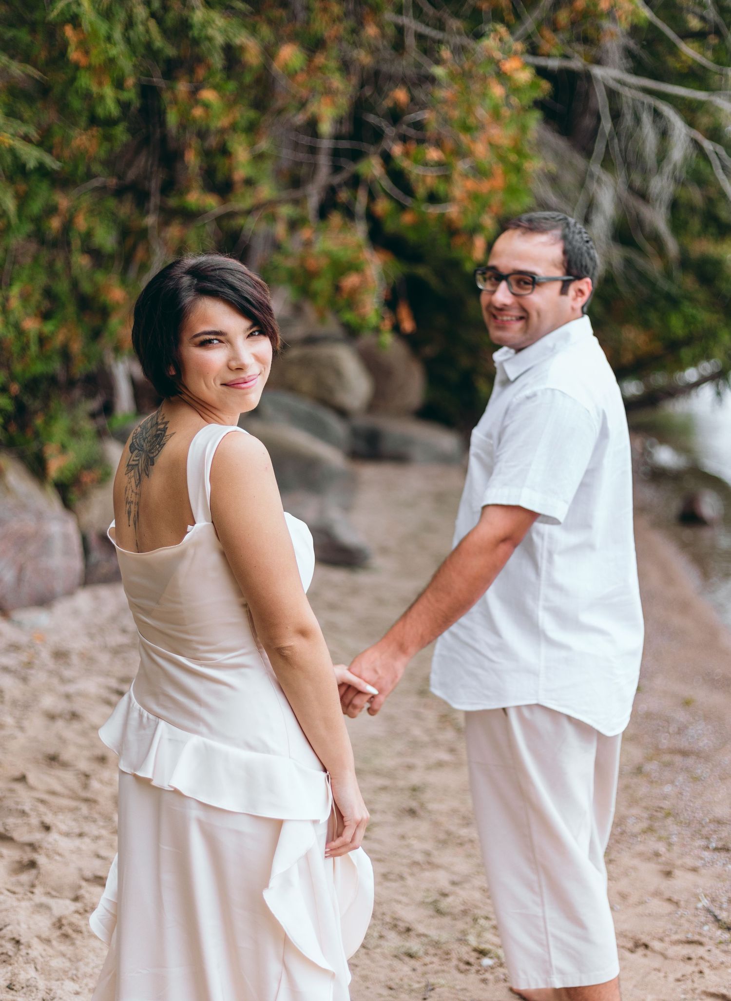 Couple in white outfits holding hands on beach with autumn foliage backdrop.
