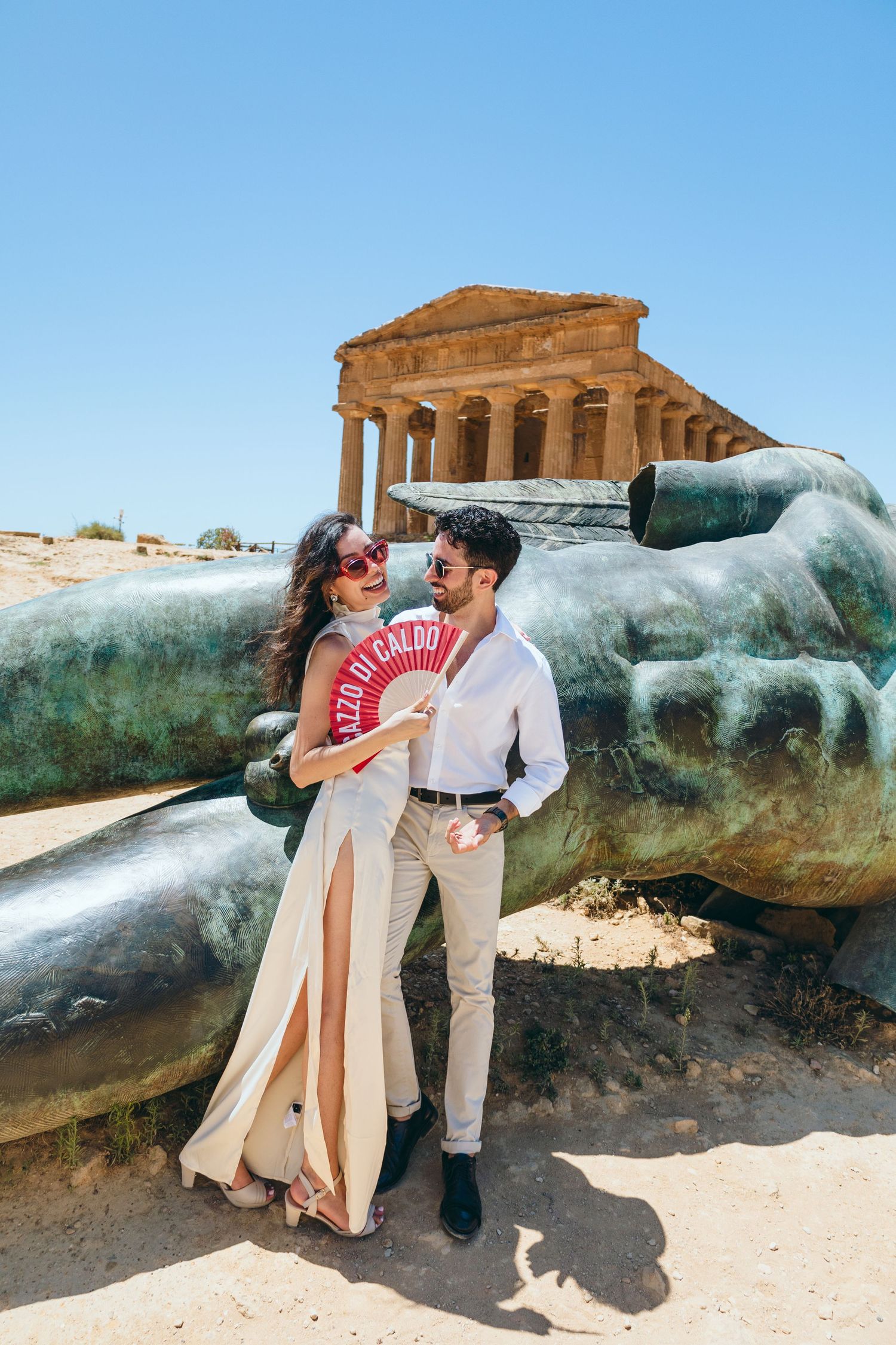 Couple in white outfits share romantic moment near fallen statue at Greek temple ruins.