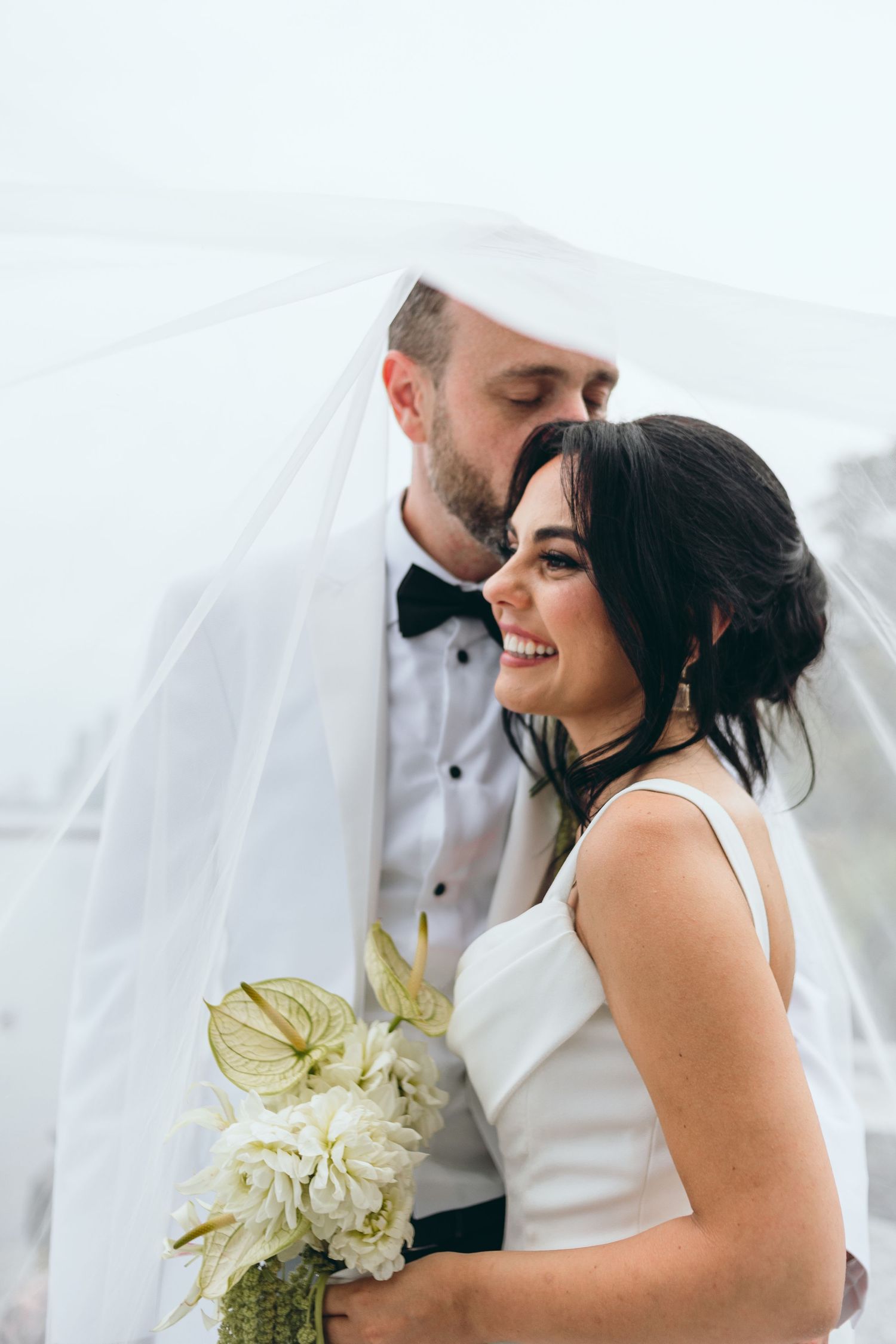 Intimate wedding moment captured under flowing white veil against bright sky.