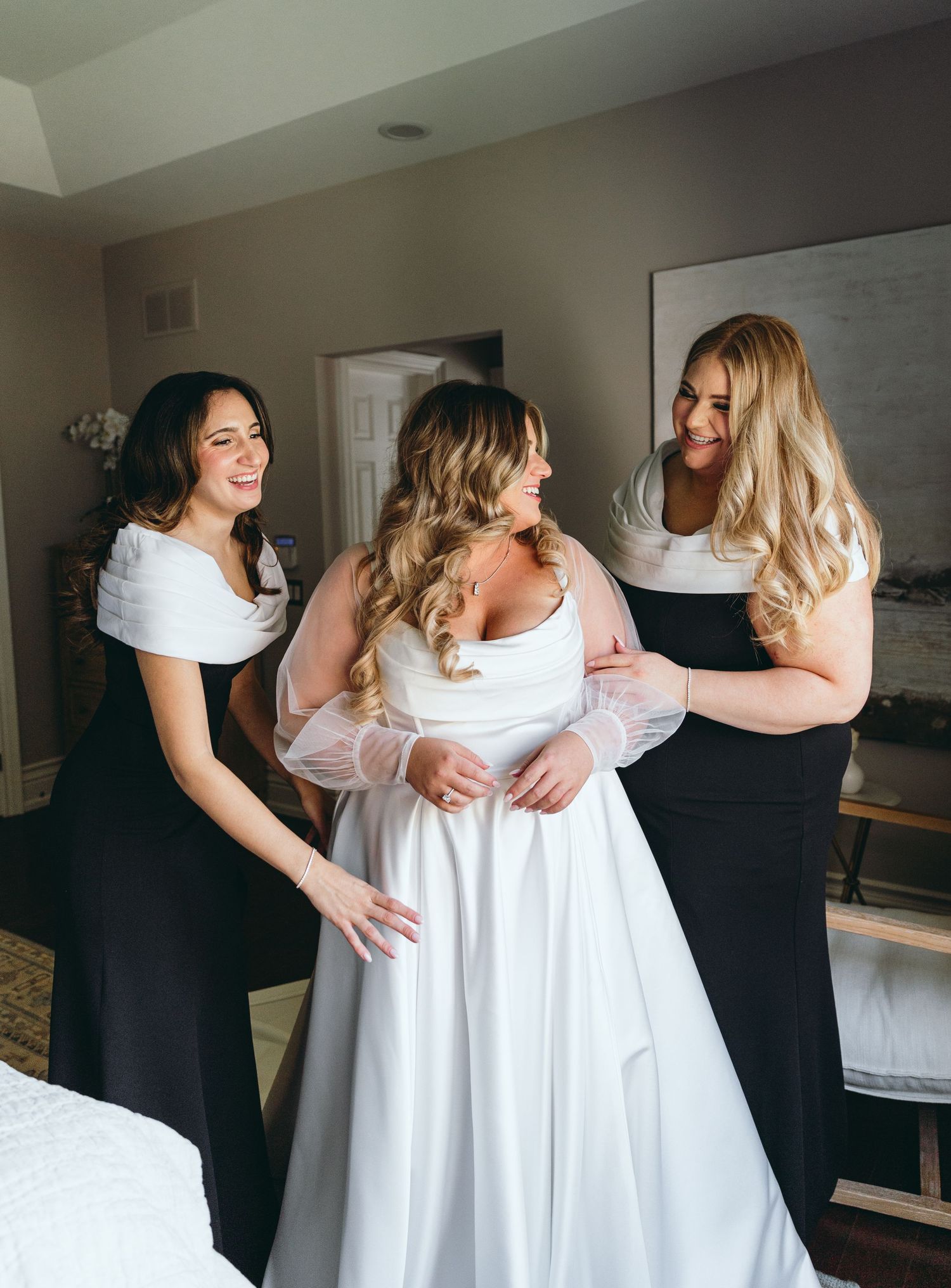 Bridesmaids in black dresses help adjust the bride's white gown during wedding preparations.