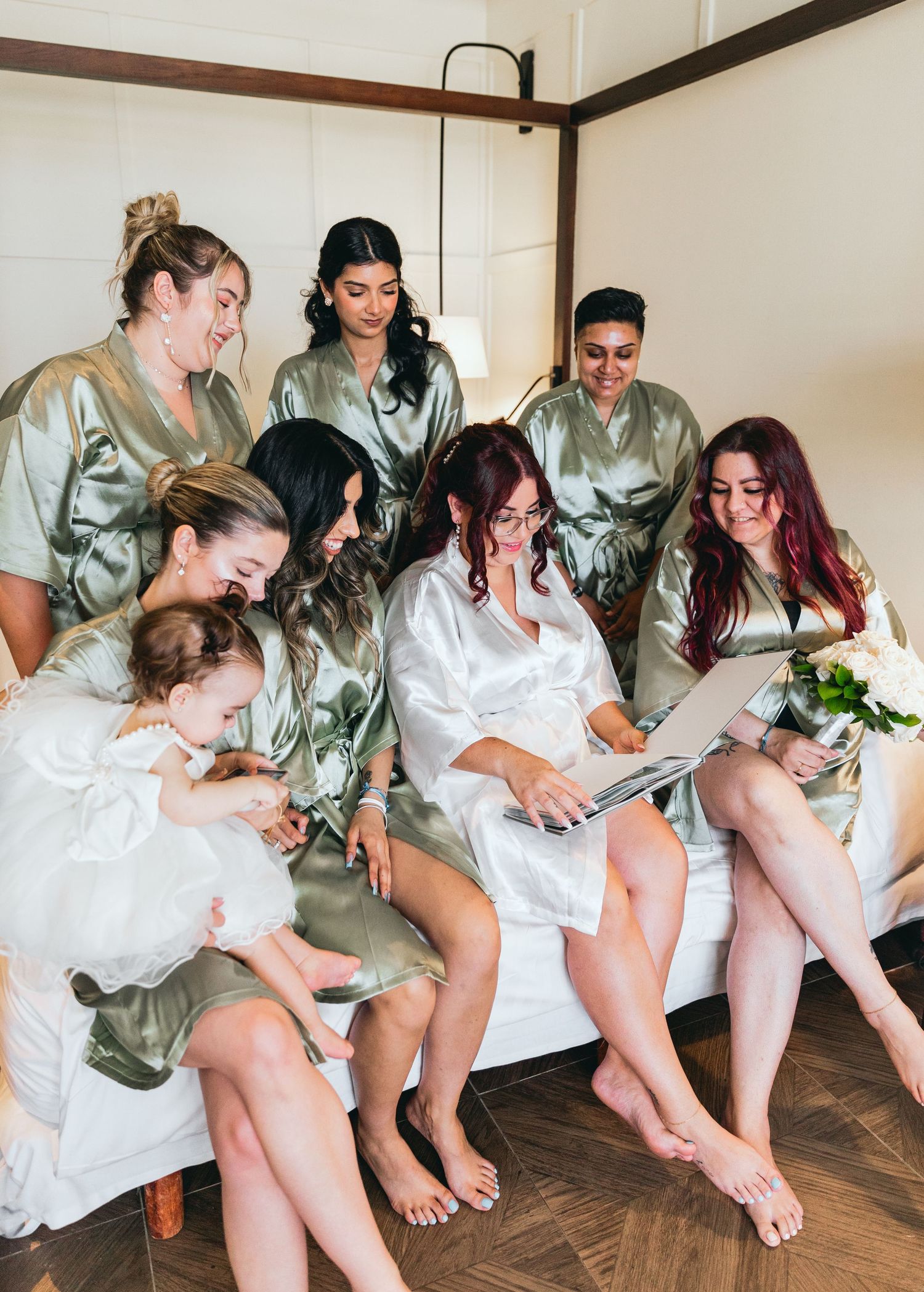 Bridesmaids in matching silk robes sit in a row on a bed during pre-wedding preparations.