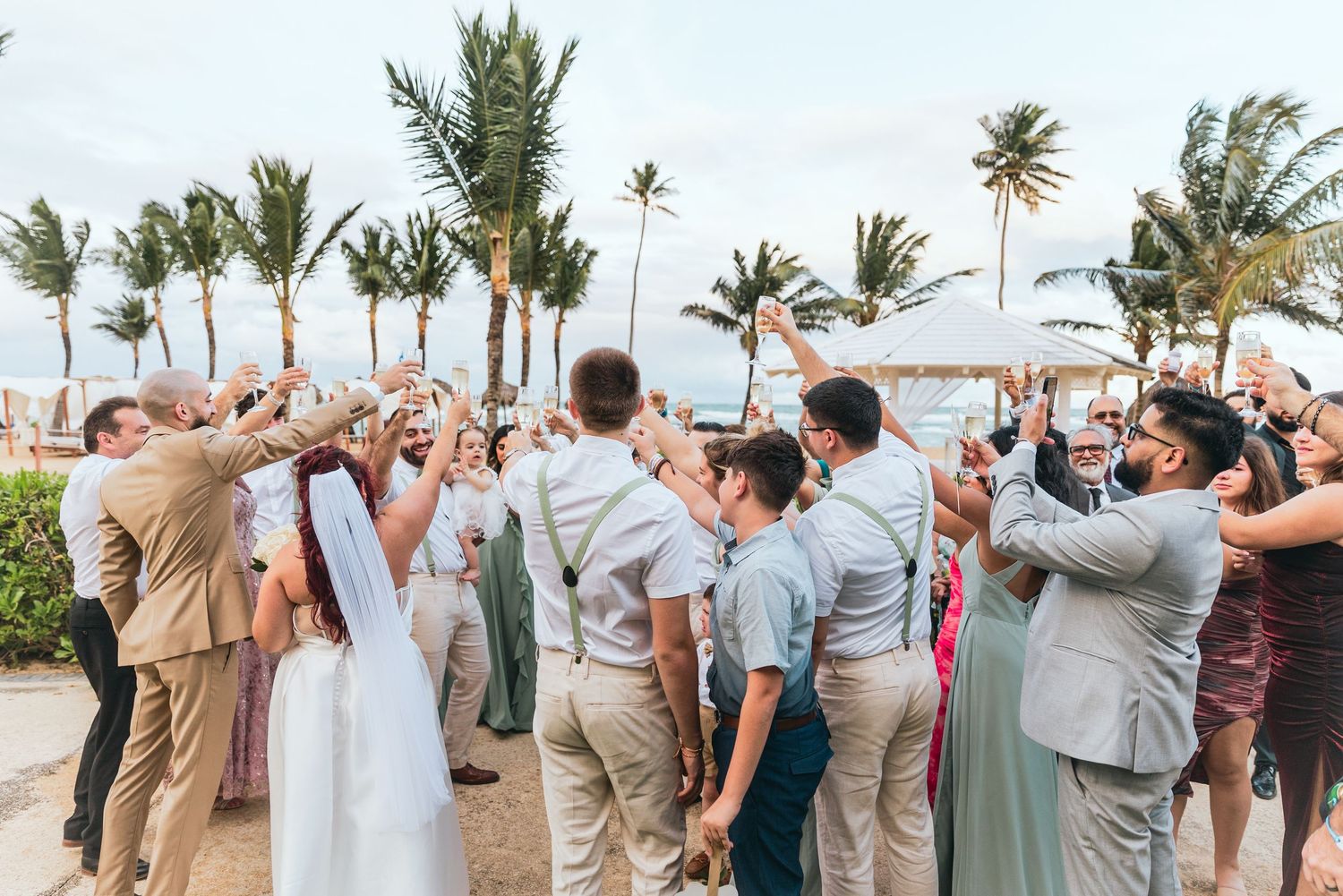 Wedding guests celebrate with raised arms against a backdrop of swaying palm trees.