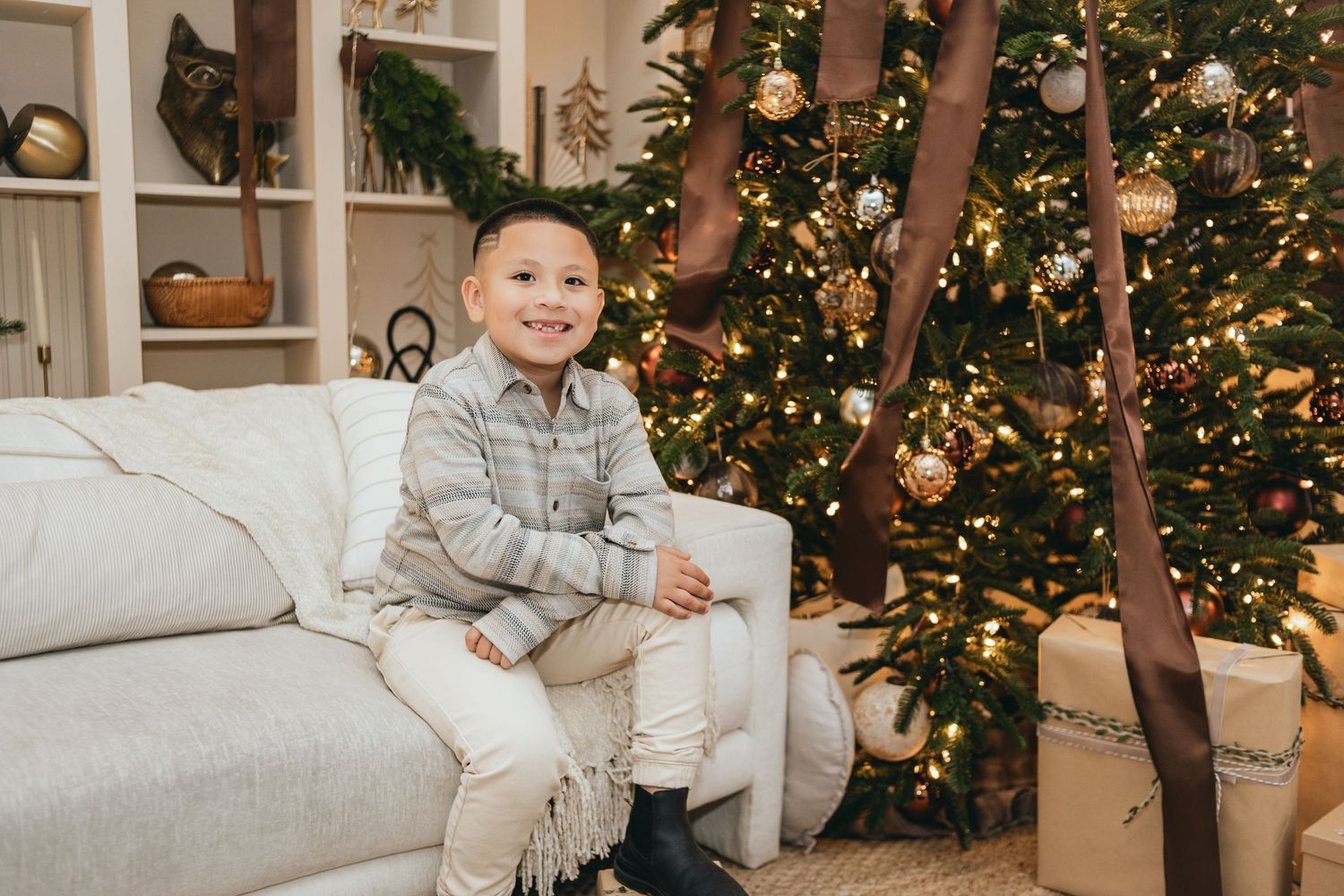 A young boy smiles on a white couch beside a decorated Christmas tree with gifts and warm lights.