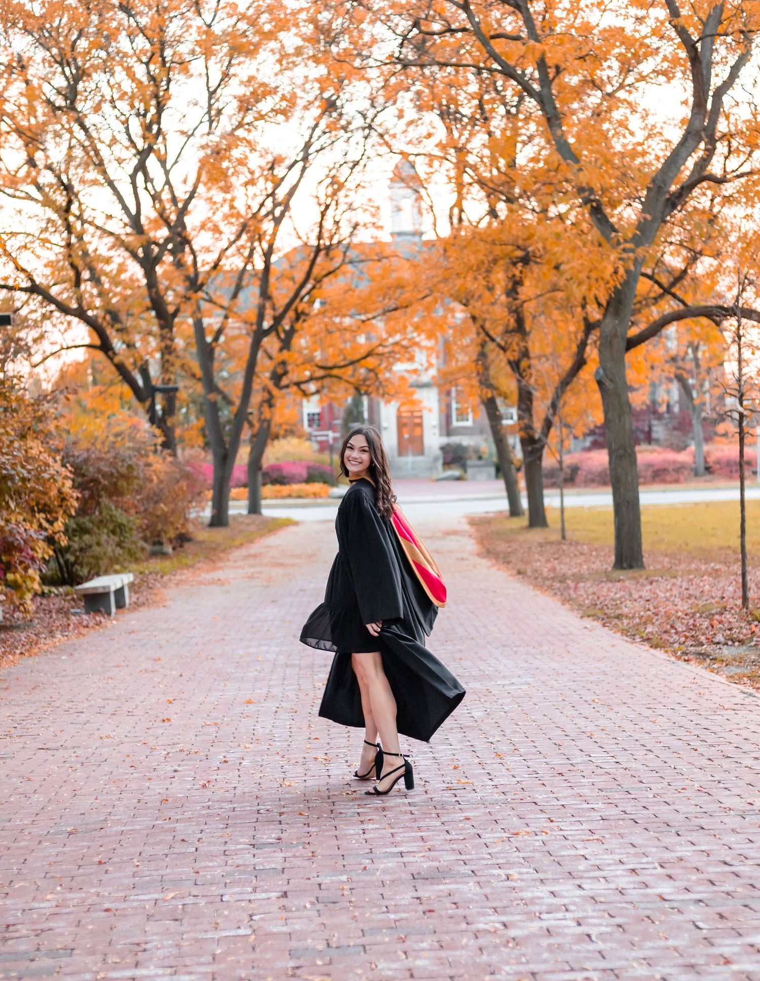 A figure in a black coat walks along a brick path surrounded by autumn trees with vibrant orange leaves.