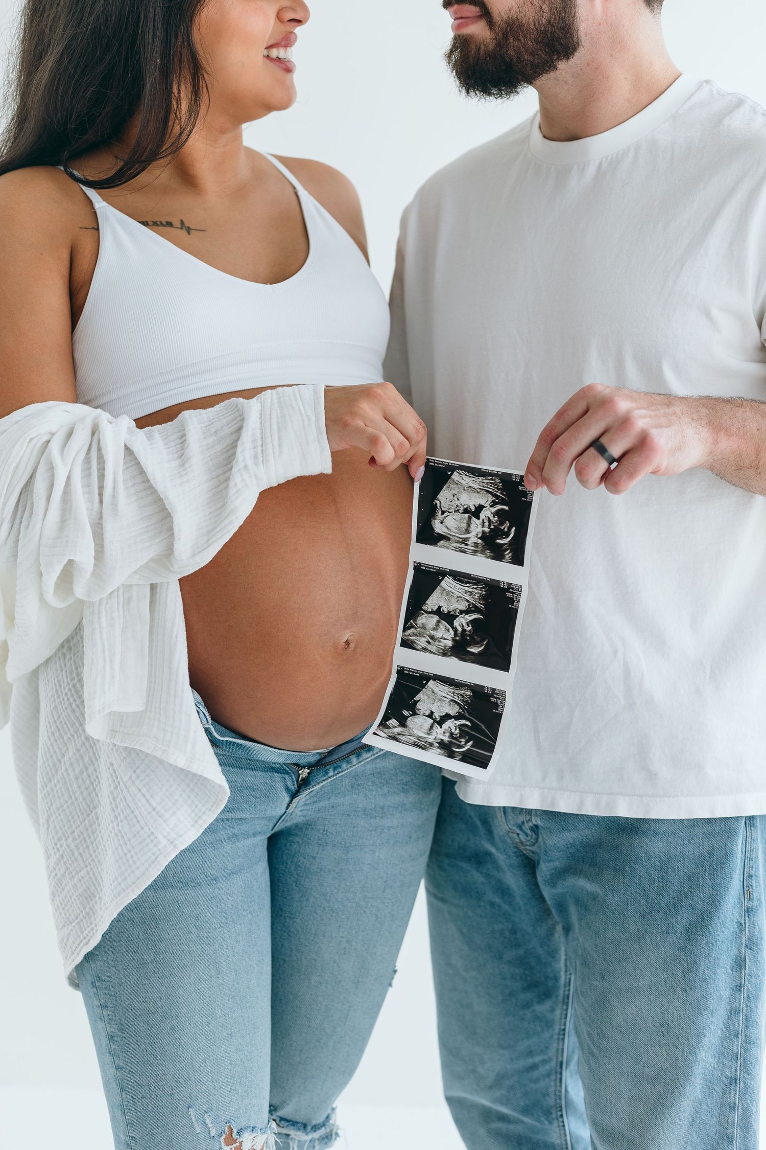 Couple holding ultrasound photos next to a pregnant belly in casual attire.